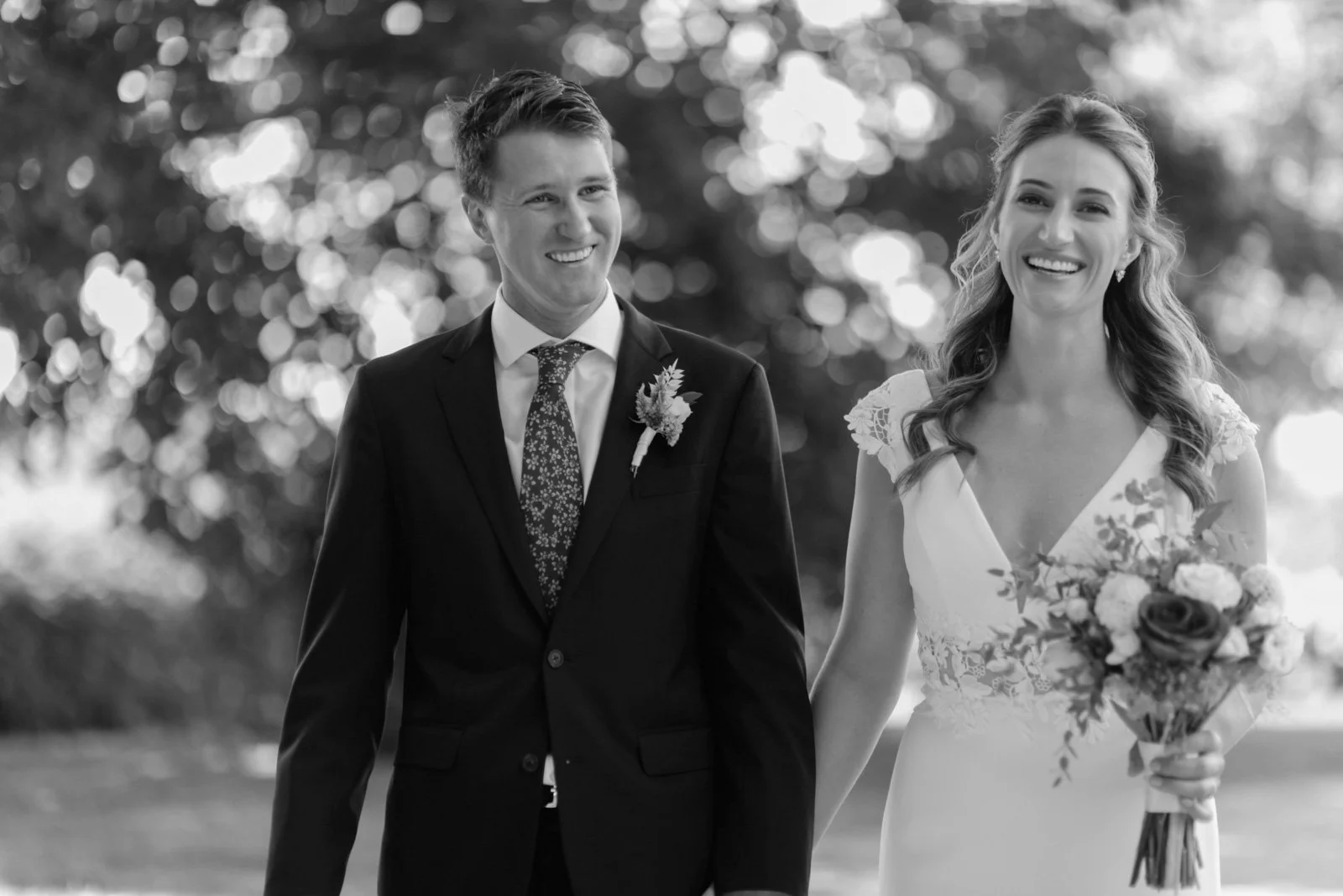 A black and white photo of a smiling bride and groom walking outdoors. The bride is holding a bouquet of flowers and wearing a wedding dress. The groom is dressed in a suit with a floral tie and boutonniere. They are holding hands and appear happy.