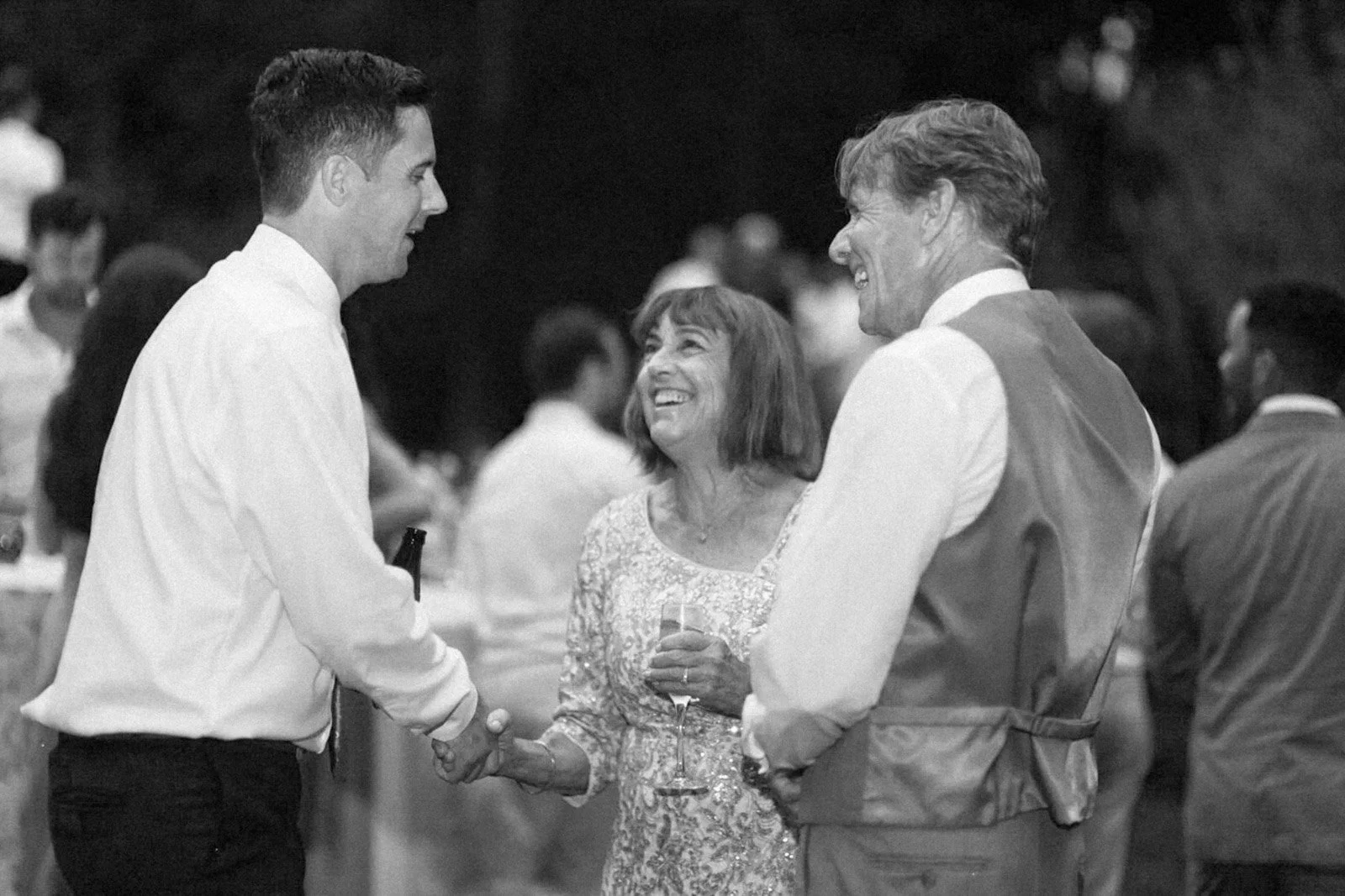 Three people smiling and talking at an outdoor event during the evening. A young man on the left is holding hands with an older woman in the middle, who is holding a glass of wine. The man on the right is looking at the woman, all are smiling, with b