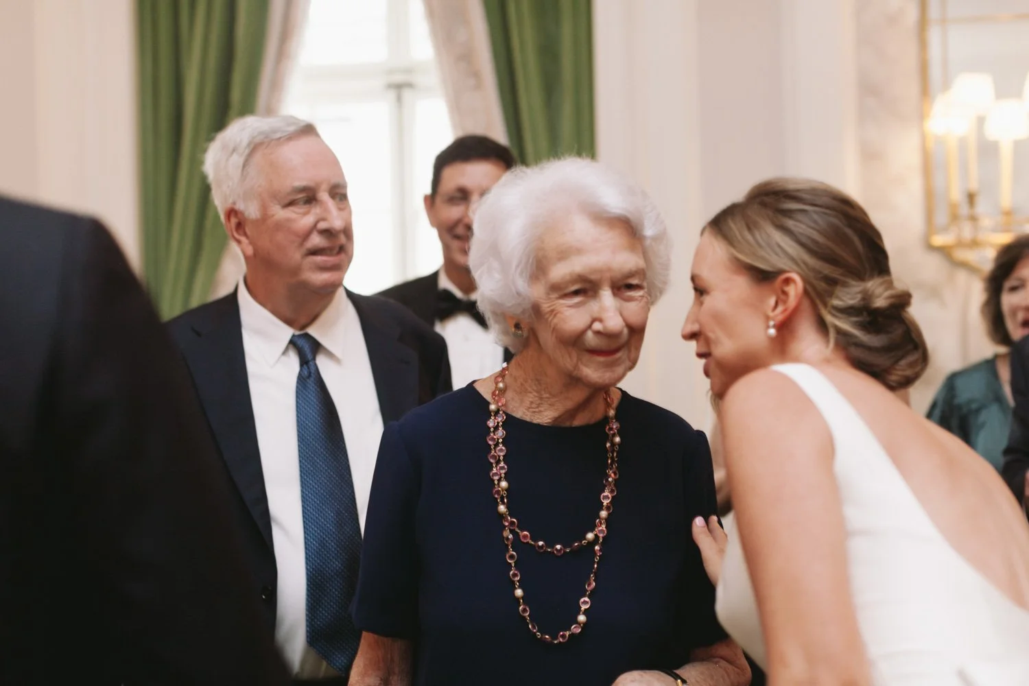 An elderly woman and a young woman are leaning towards each other at a formal gathering, with three men in the background.