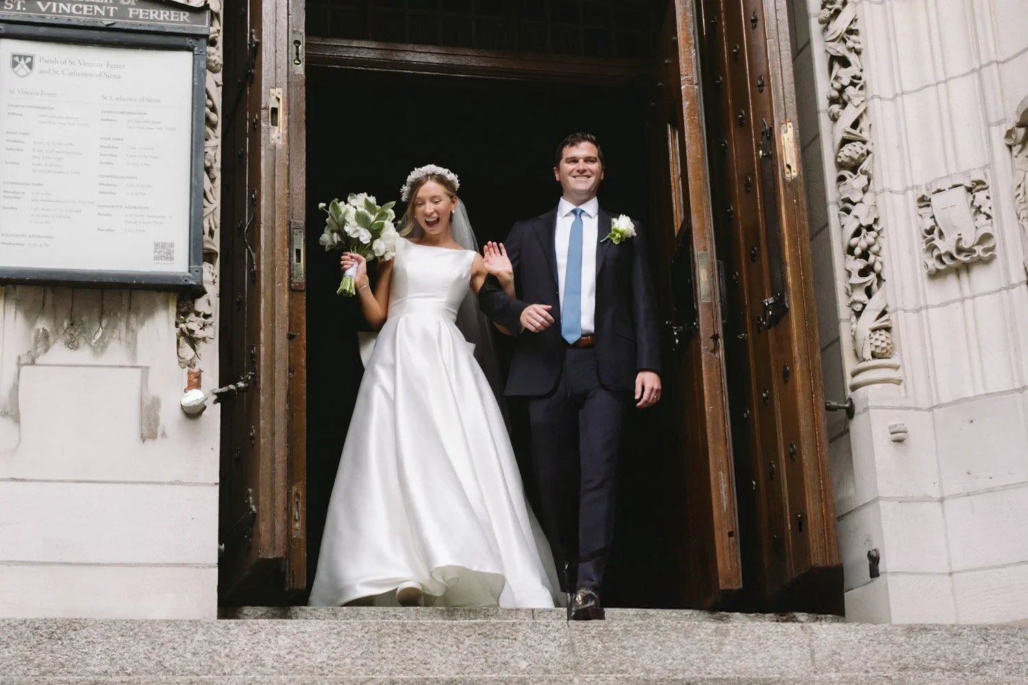 A newlywed couple exiting a church, the bride in a white gown holding a bouquet and the groom in a dark suit with a blue tie, both smiling happily.