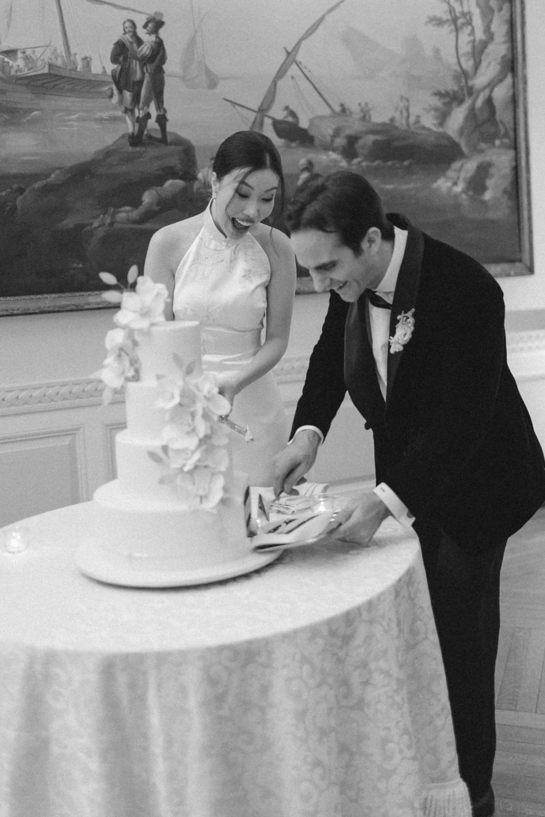 A black-and-white photo of a bride and groom slicing a wedding cake together at their wedding reception, with a large painting in the background.