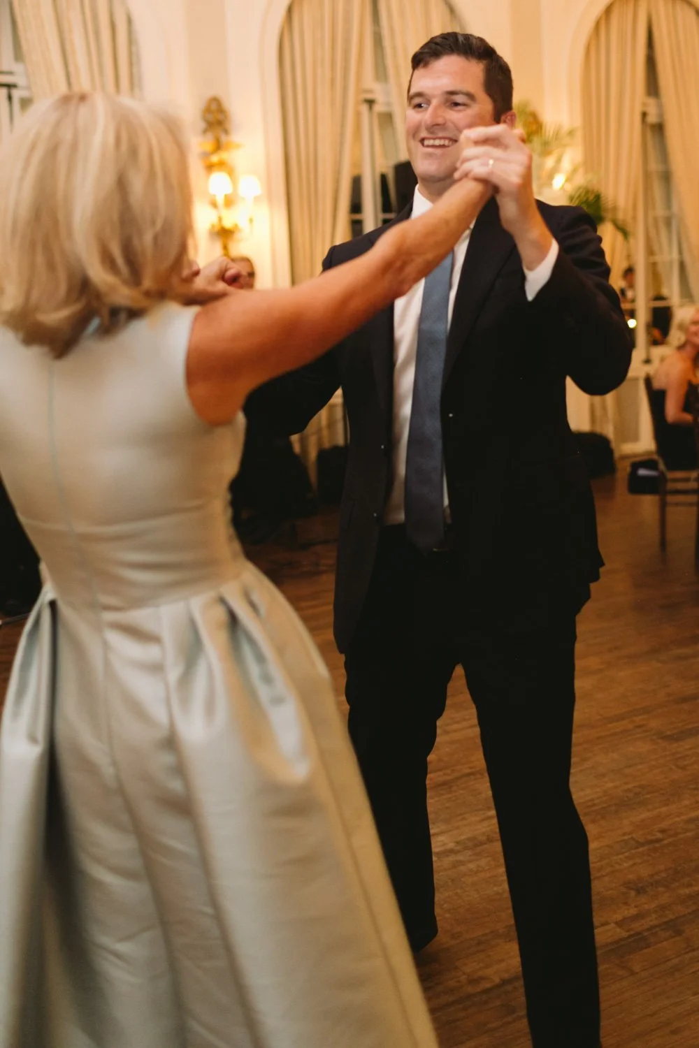 A man and woman dancing together at a formal event in a decorated room with cream-colored curtains, warm lighting, and other people in the background.