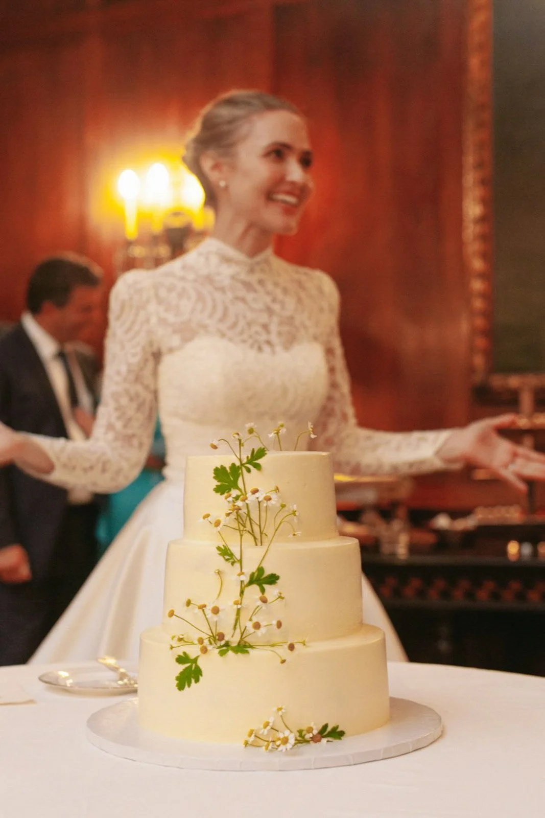 A wedding cake with three tiers decorated with small white flowers and green leaves, placed on a white tablecloth. In the background, a woman in a white lace wedding dress is smiling, with warm lighting and wood-paneled walls.