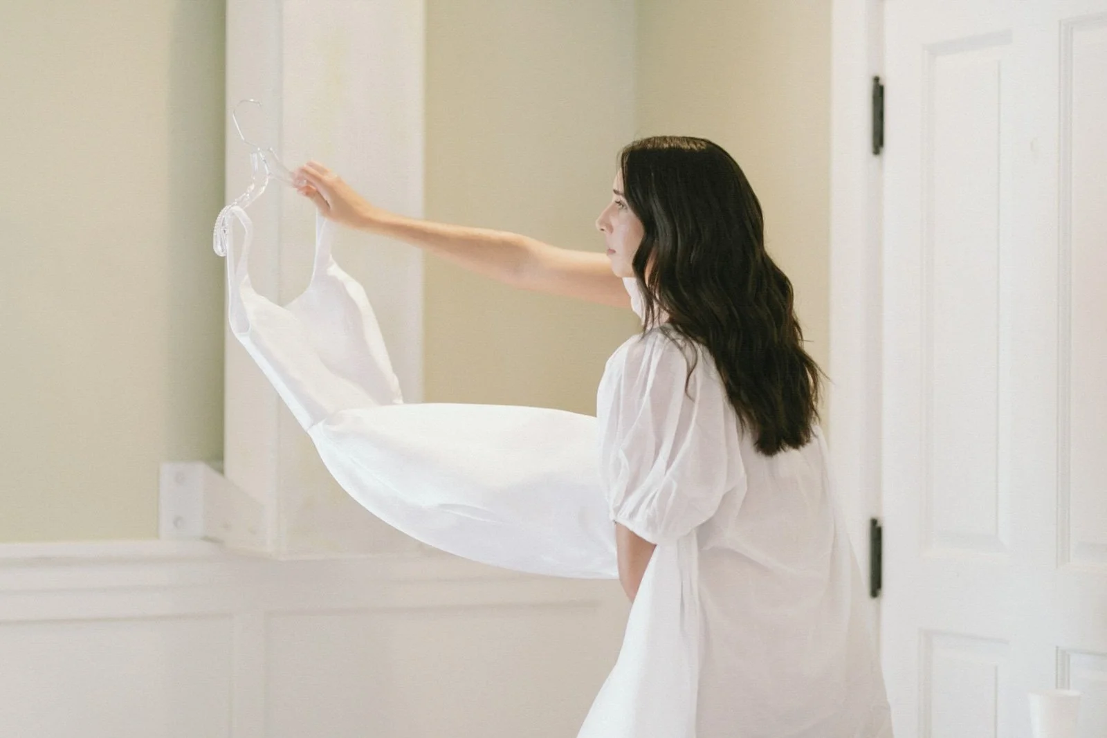 A woman with dark wavy hair in a white dress hanging a white slip dress on a hanger in a room with white walls and a door.