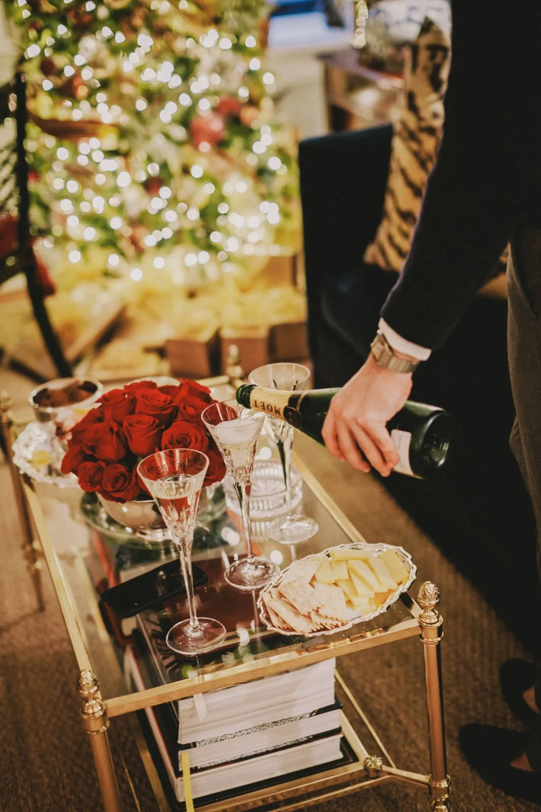 A person pouring champagne into glasses on a table decorated with a bouquet of red roses, cheese, and charcuterie, with a Christmas tree in the background.