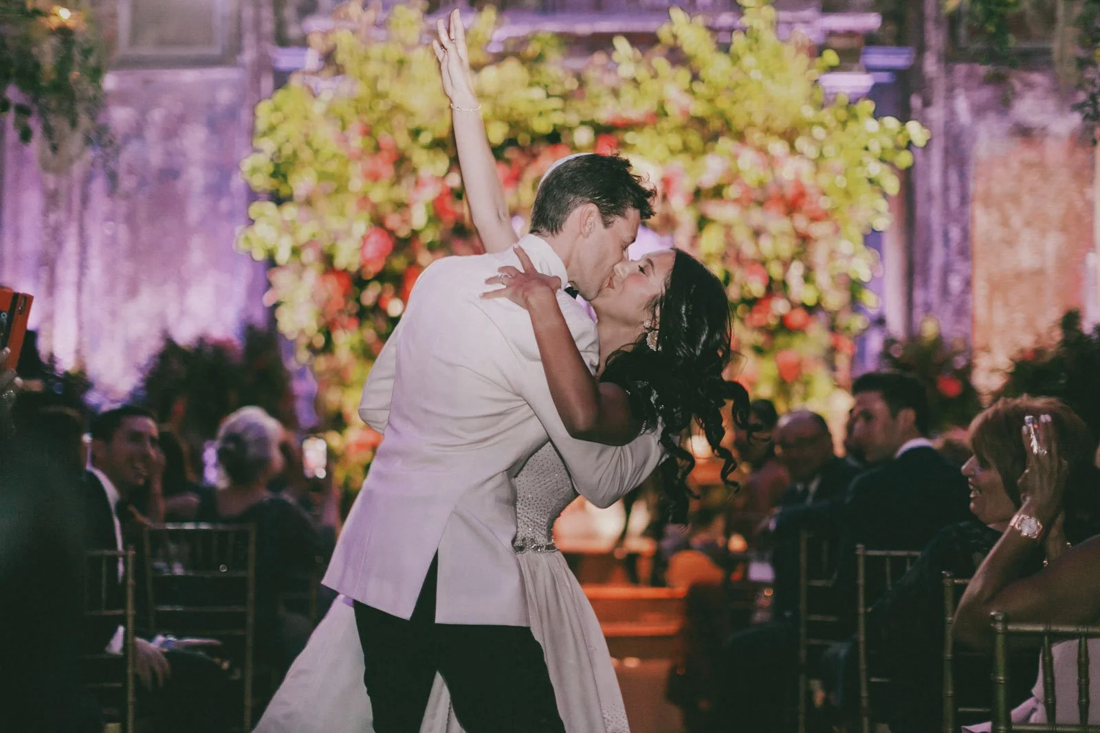 A couple dressed in wedding attire shares a kiss during their wedding reception, with a large floral arrangement and guests seated at tables in the background.