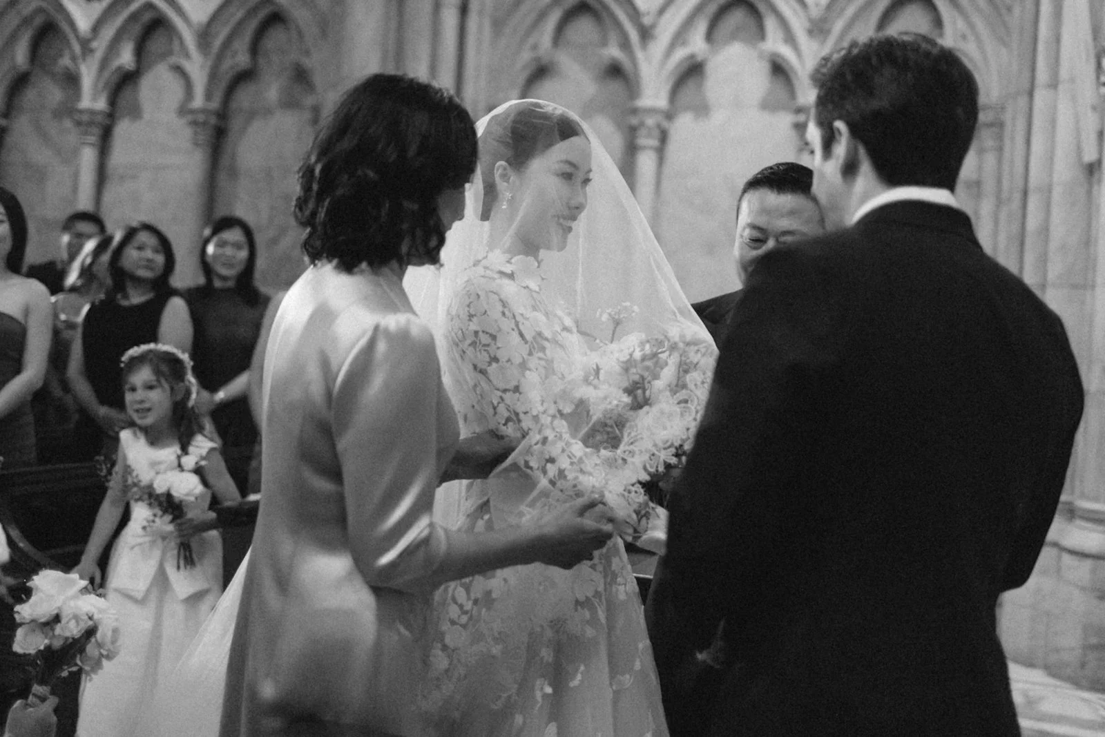A black-and-white photograph of a wedding ceremony. The bride, in a lace wedding dress and veil, is holding hands with the groom, who is dressed in a suit. They are standing in a church with ornate arches, surrounded by an officiant, a woman in a lig