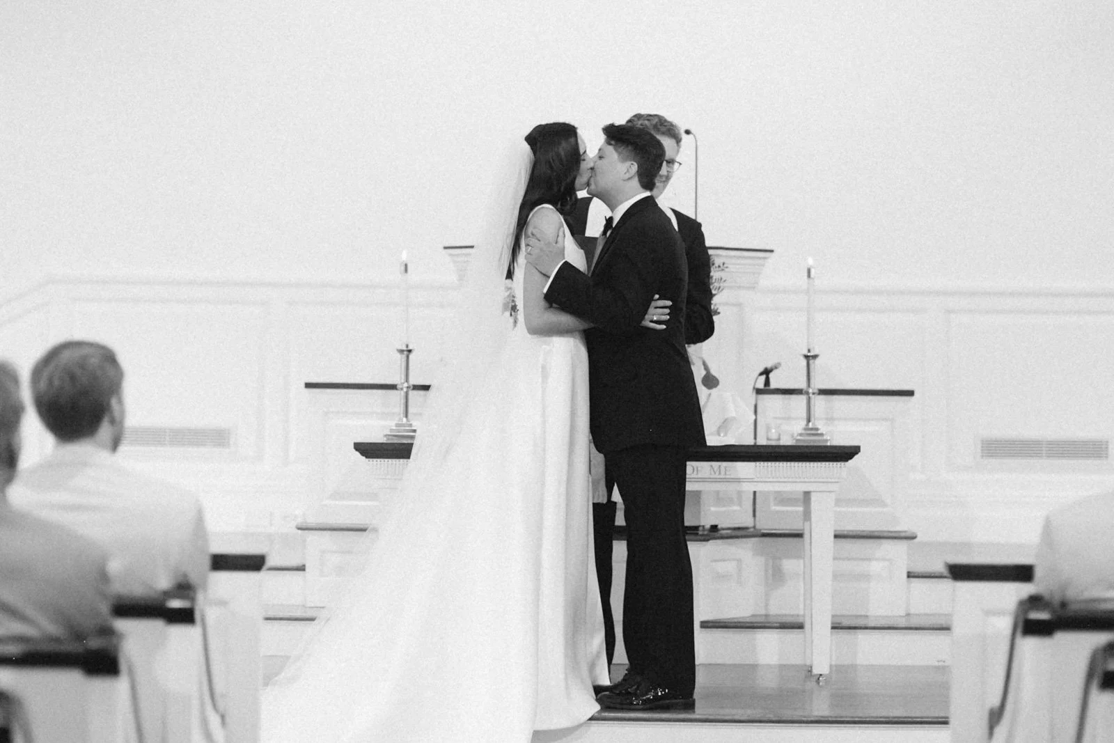 A couple in wedding attire kissing during a wedding ceremony, with an officiant standing behind them and guests seated in the foreground.