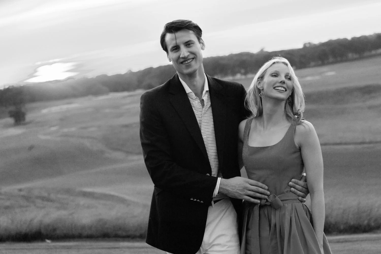 A smiling couple stands outdoors on a grassy field, the man in a blazer and the woman in a sleeveless dress, holding each other as the sun sets in the background.