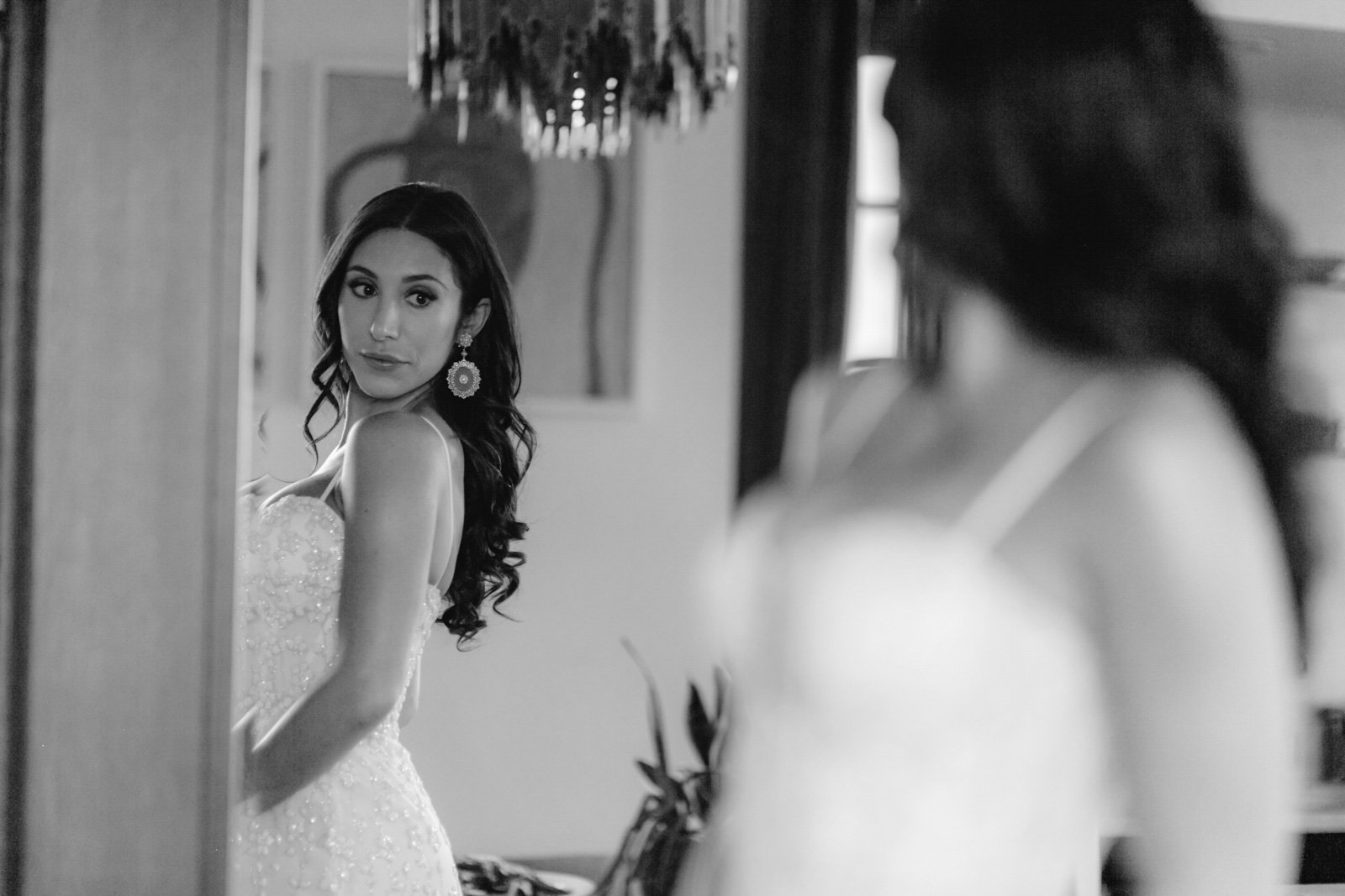 A woman in a wedding dress looking at herself in a mirror, with another woman's reflection partially visible.