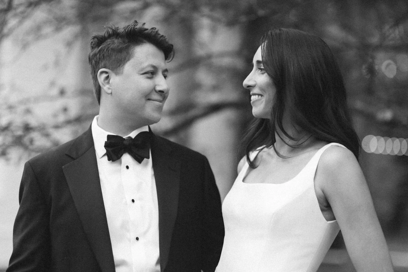 A couple looking at each other and smiling, dressed in formal attire, standing outdoors.