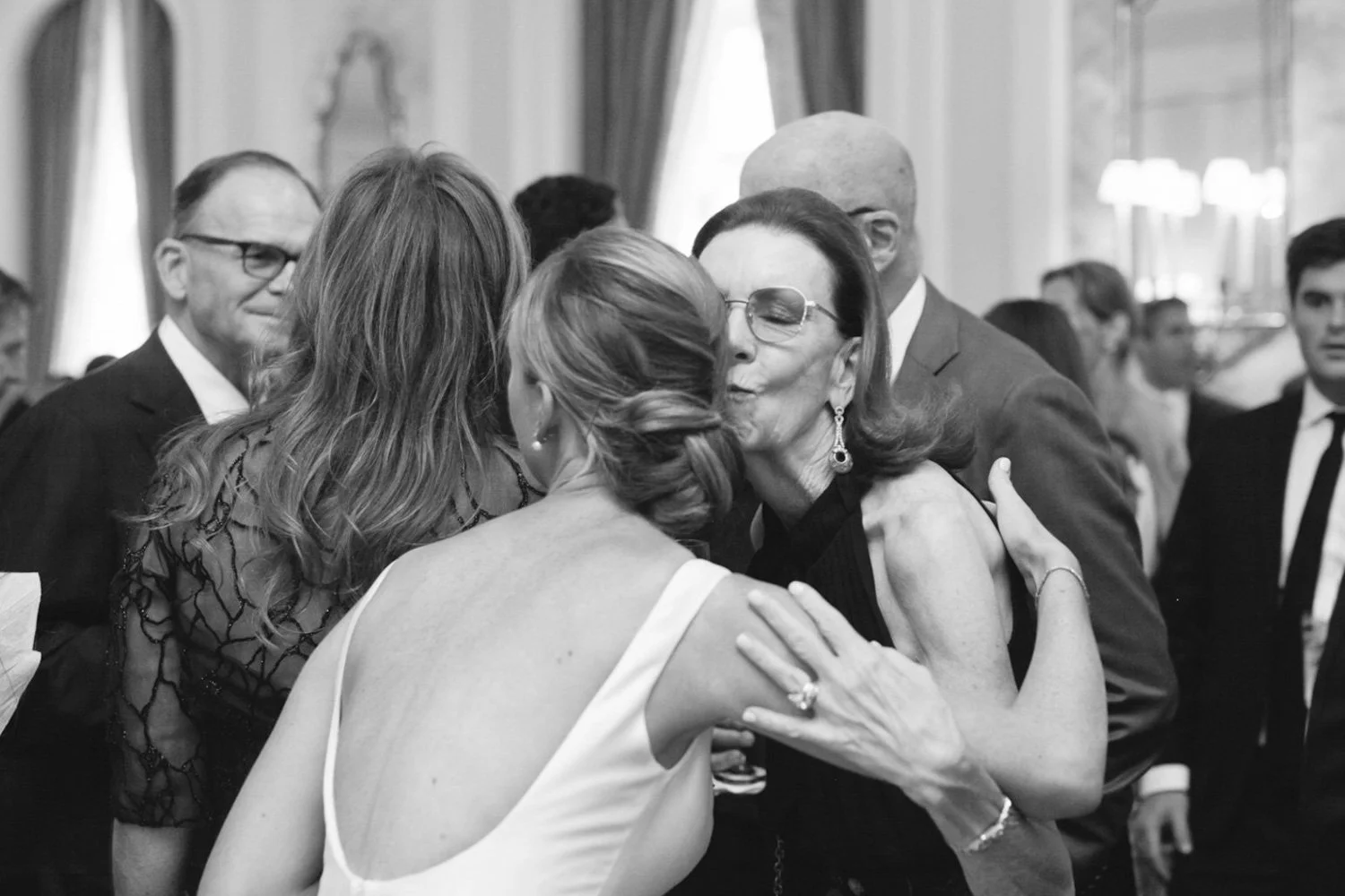 A woman in a white dress and a woman in a black dress embracing at a formal event, surrounded by other formally dressed guests in a luxurious room.
