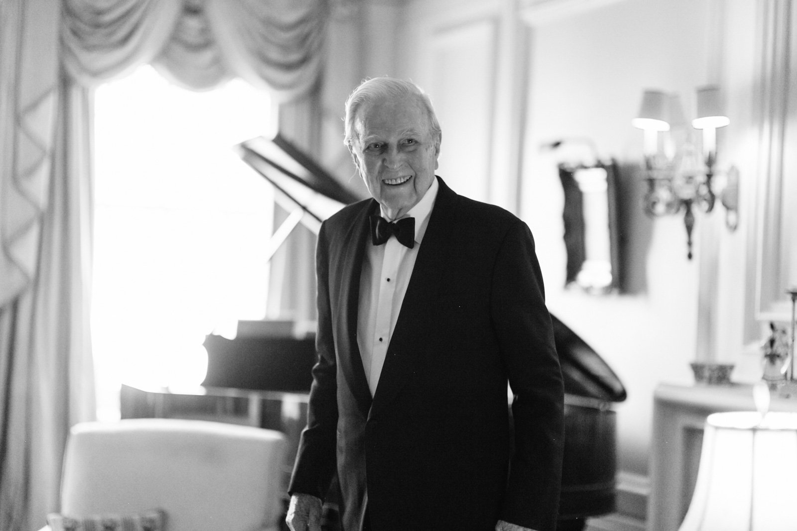 An elderly man in a tuxedo smiling in a luxurious room with a grand piano and elegant decor.