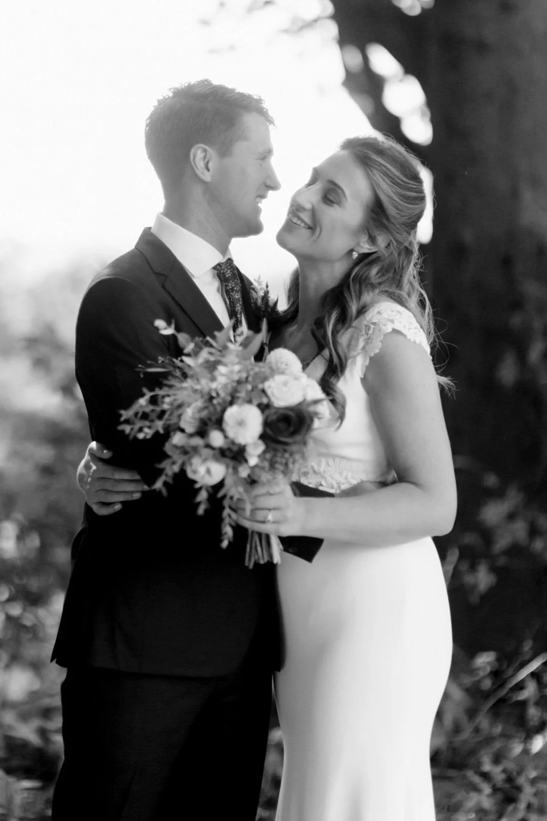 A black and white photo of a bride and groom smiling and looking at each other outdoors, with the bride holding a bouquet of flowers.