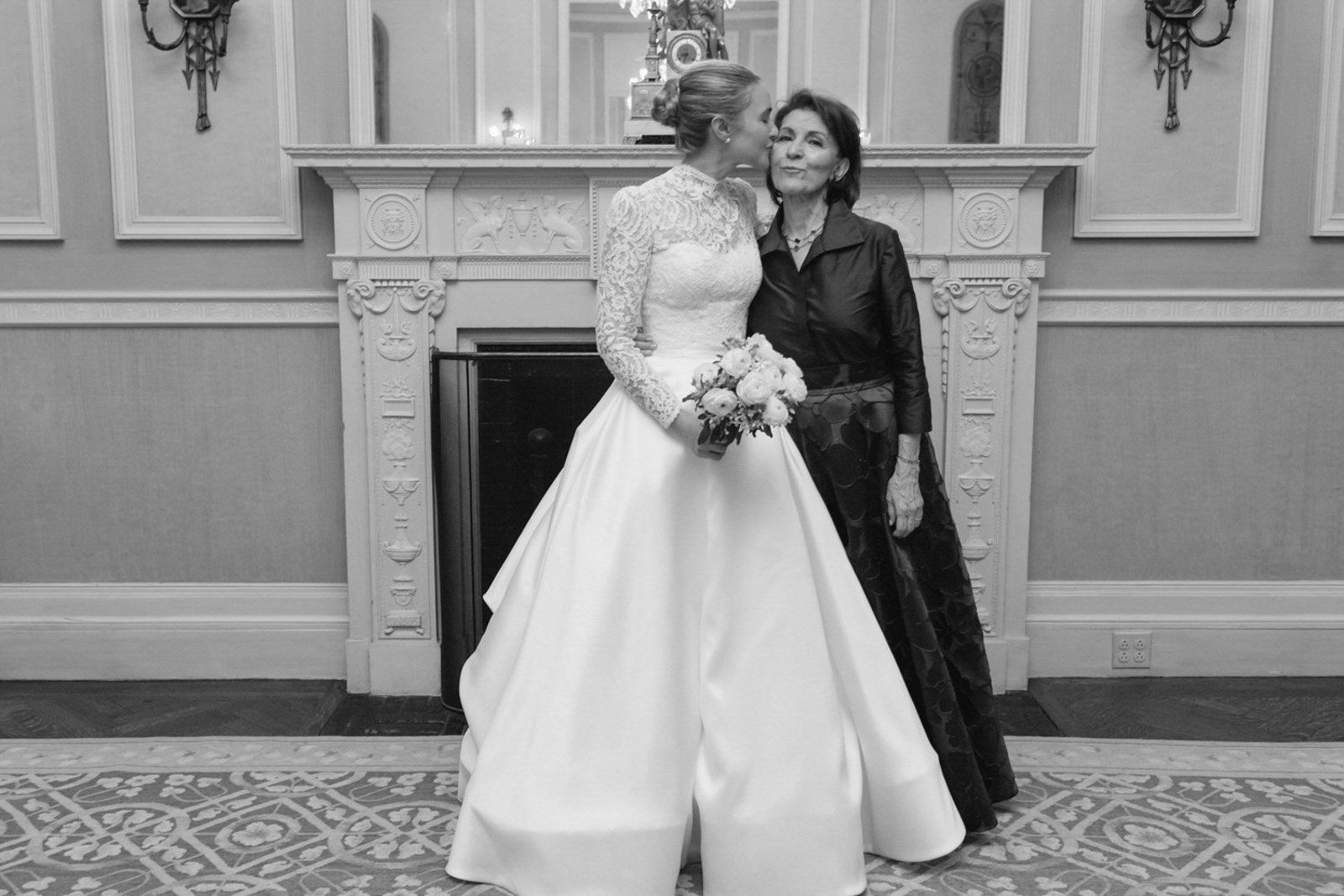 A bride in a lace long-sleeve wedding gown holding a bouquet, kissing an older woman in a dark patterned dress, standing in front of a decorative fireplace in an elegant room at The Colony Club New York.