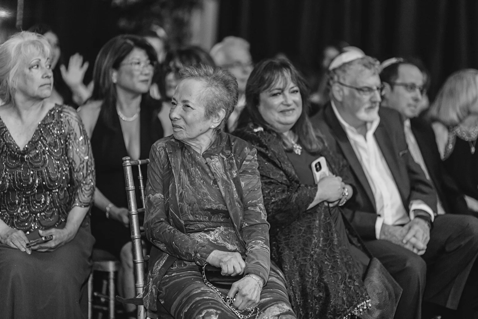 Black and white photo of a diverse group of women and a man sitting in rows, attending an event. They are dressed formally, with some looking at the stage and some looking at the camera.