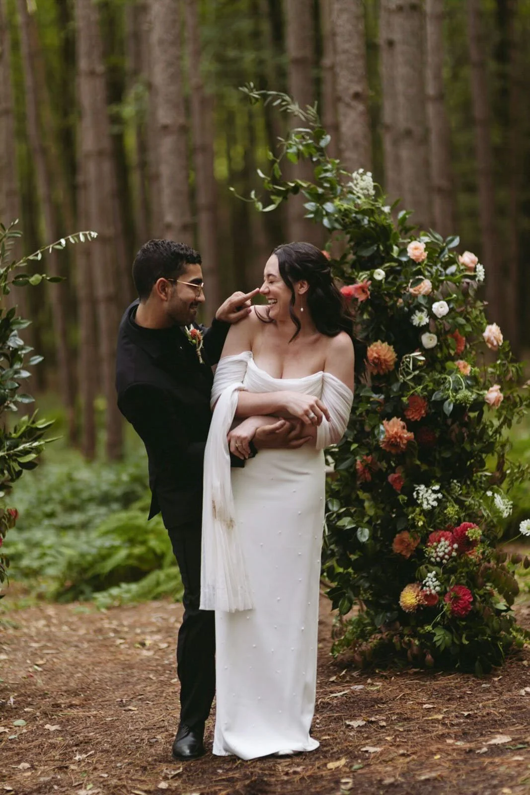 A happy couple wedding photoshoot in the woods, with the groom holding the bride who is smiling and laughing, surrounded by greenery and floral decorations.