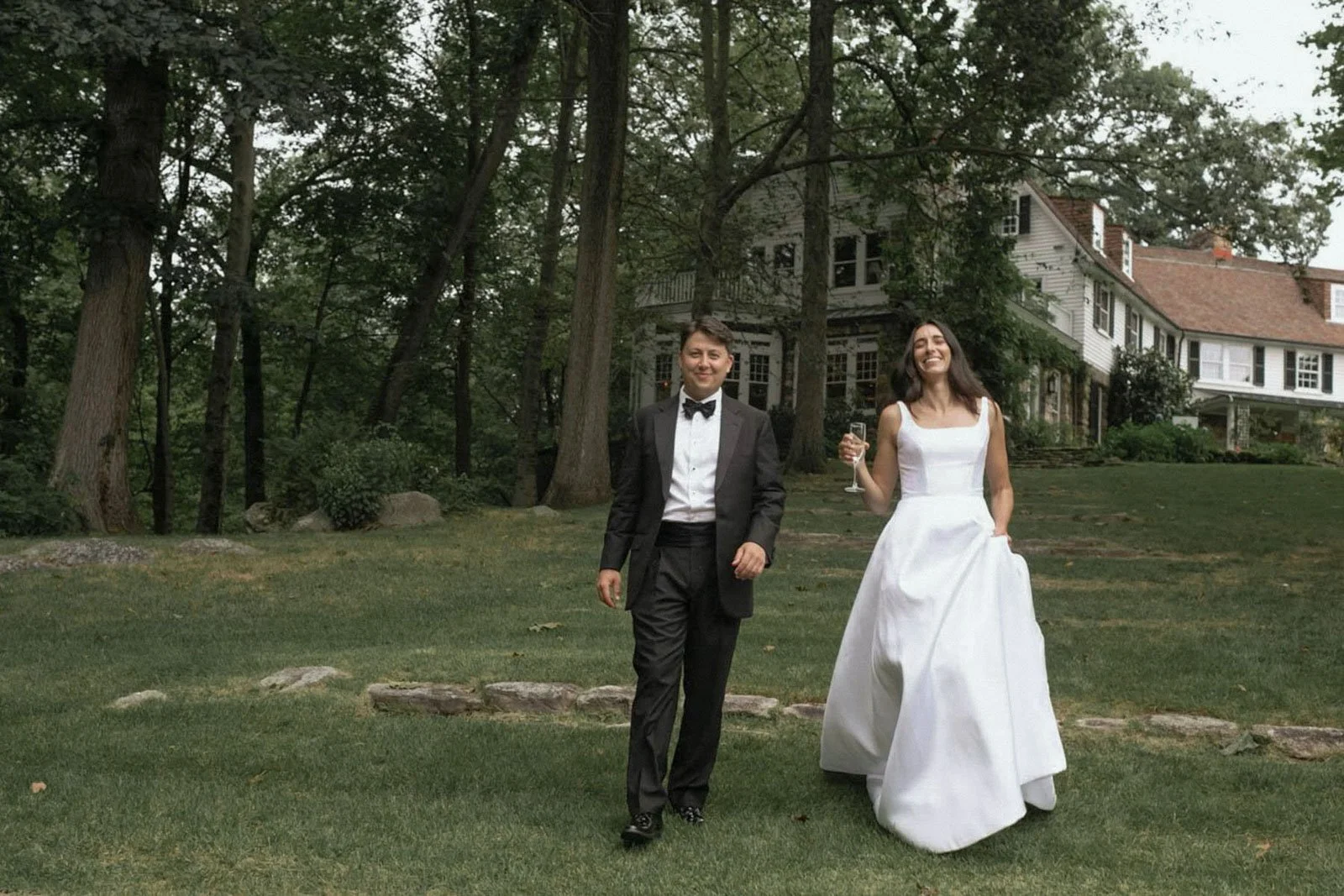 A newlywed couple walking on a grassy lawn, with the groom in a black tuxedo and bow tie and the bride in a white wedding dress holding a glass of champagne, in front of a large house and trees.