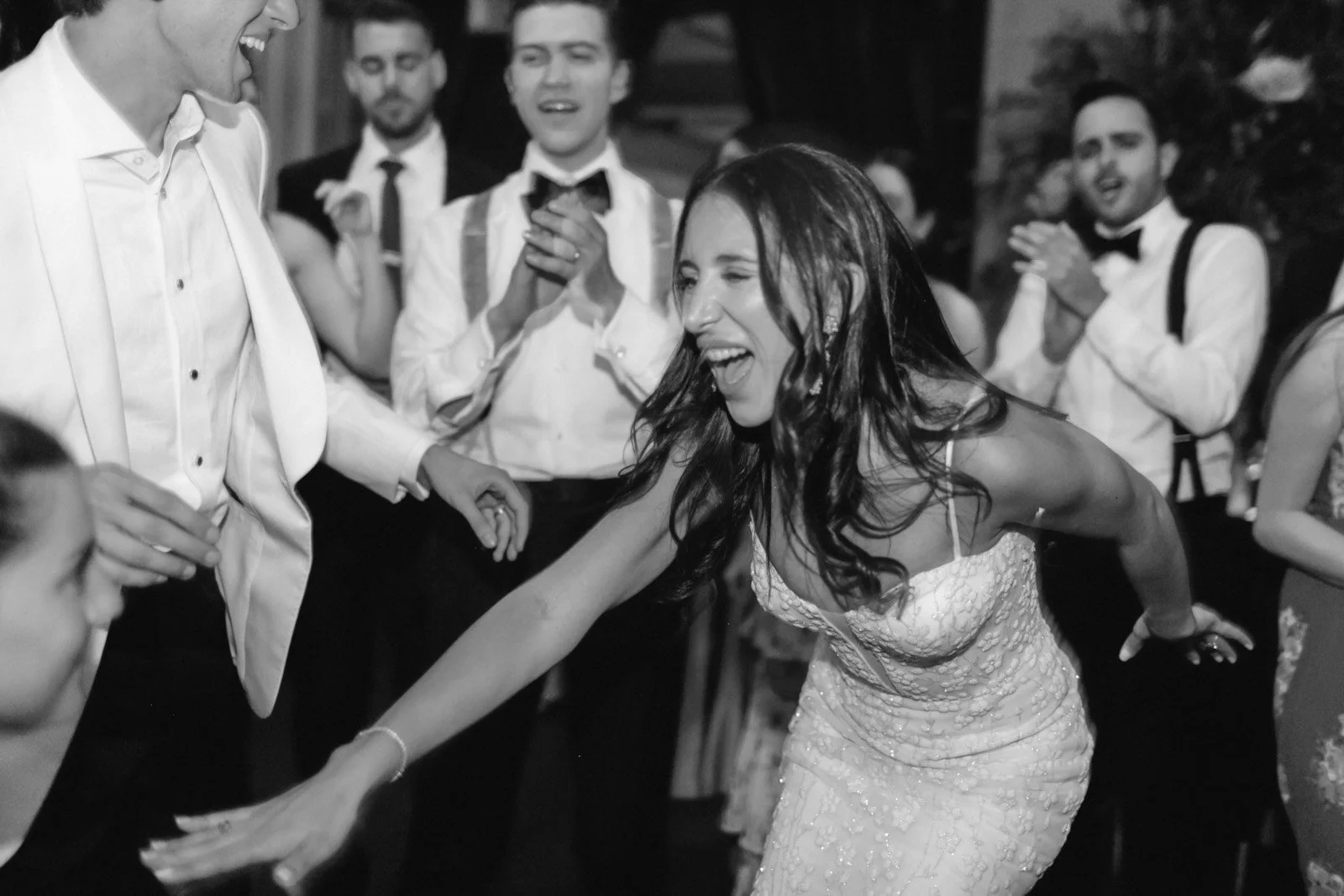A woman in a wedding dress laughing and reaching out, surrounded by men in tuxedos at a celebration or wedding reception.