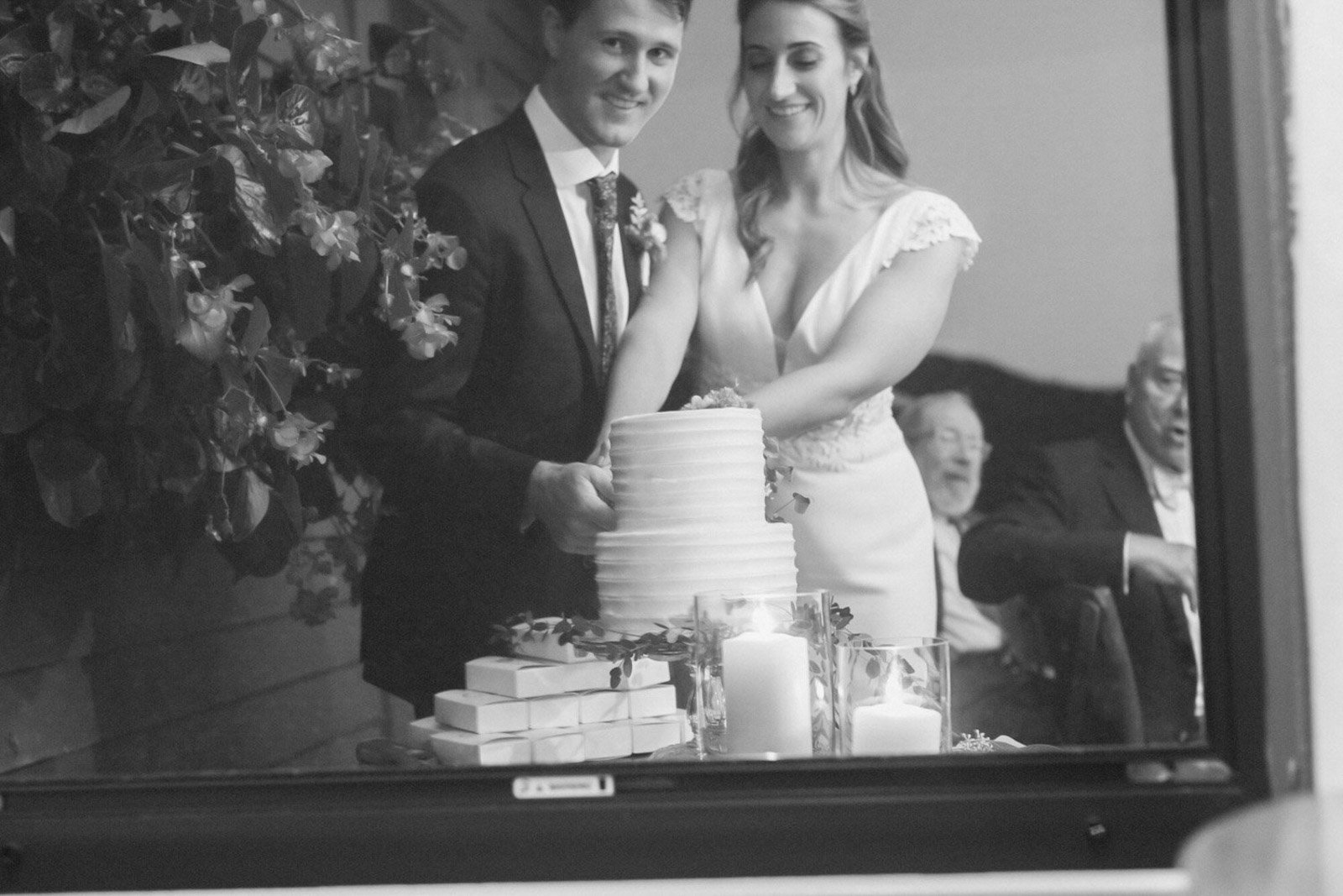 A black-and-white photograph of a bride and groom cutting their wedding cake, viewed through a window or mirror with candle decorations nearby, with three guests in the background at Court Hill in The Berkshires.