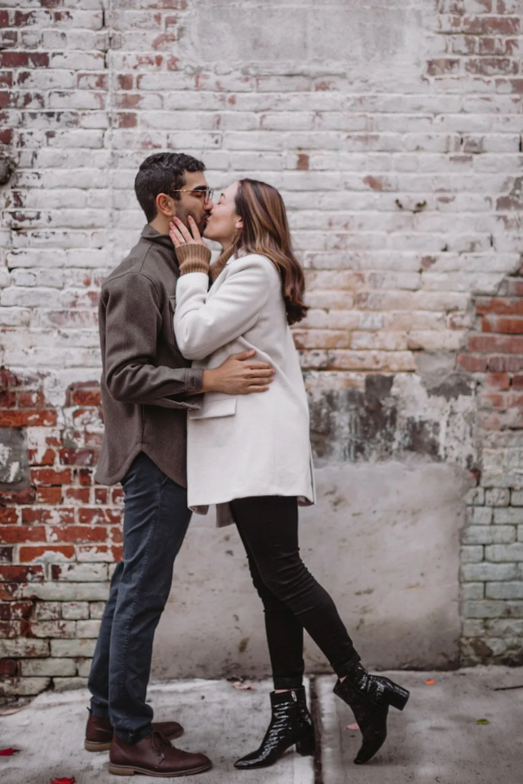 A man and woman are kissing against a weathered brick wall, the woman’s hands on the man's face and his arm around her waist, sharing an intimate moment.