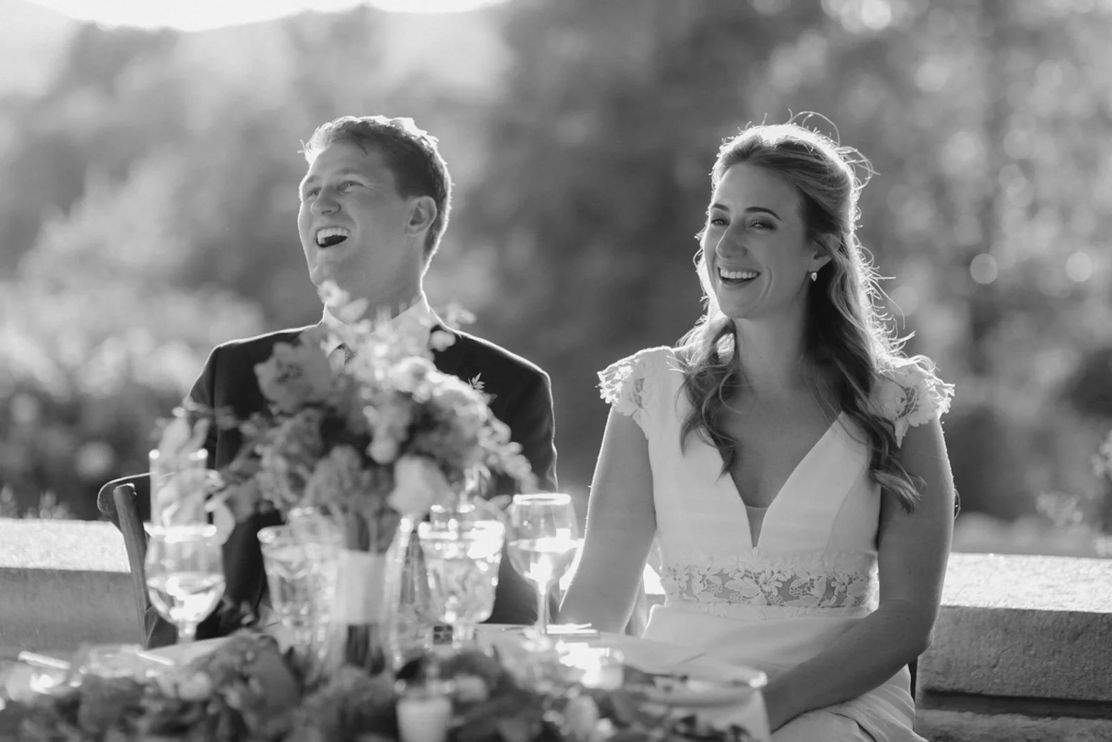 A black and white photo of a smiling man and woman seated at a table outdoors, possibly at a wedding reception, with flowers and glasses on the table, and trees in the background.