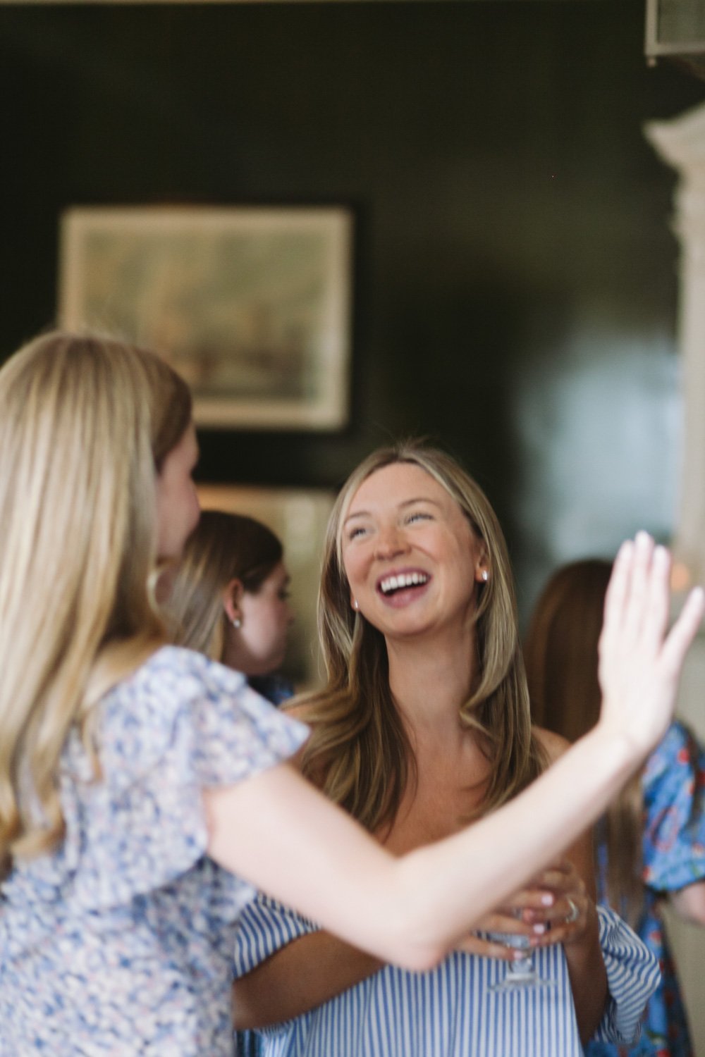 A group of women socializing indoors, smiling, with one woman gesturing with her hand as others listen, in a warm and lively setting.