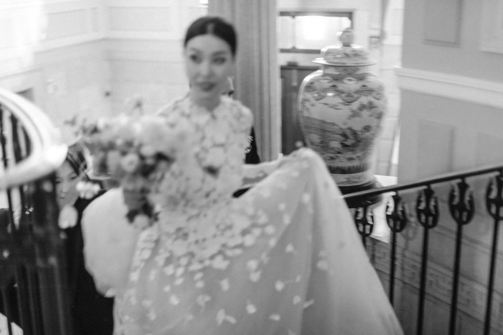 A woman in floral dress standing behind a metal railing, holding a piece of cake, with a large decorative vase in the background.