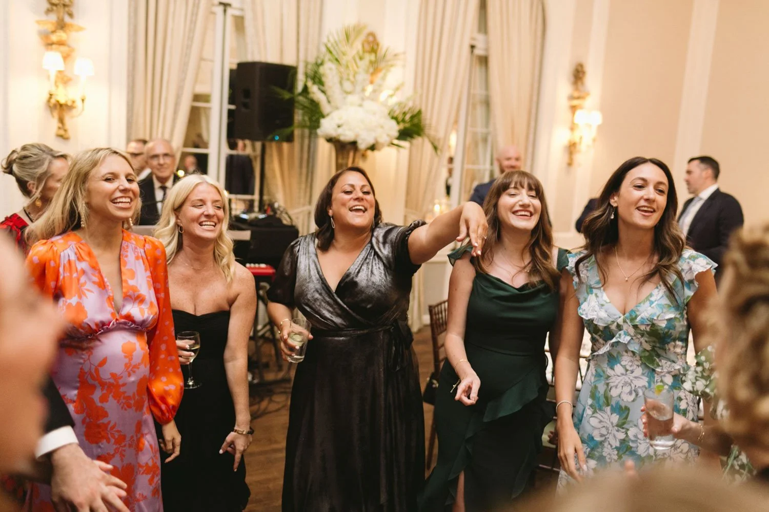 Group of women at a party or celebration, smiling and gesturing, with drinks in hand, in an elegant indoor setting with curtains and floral centerpieces.