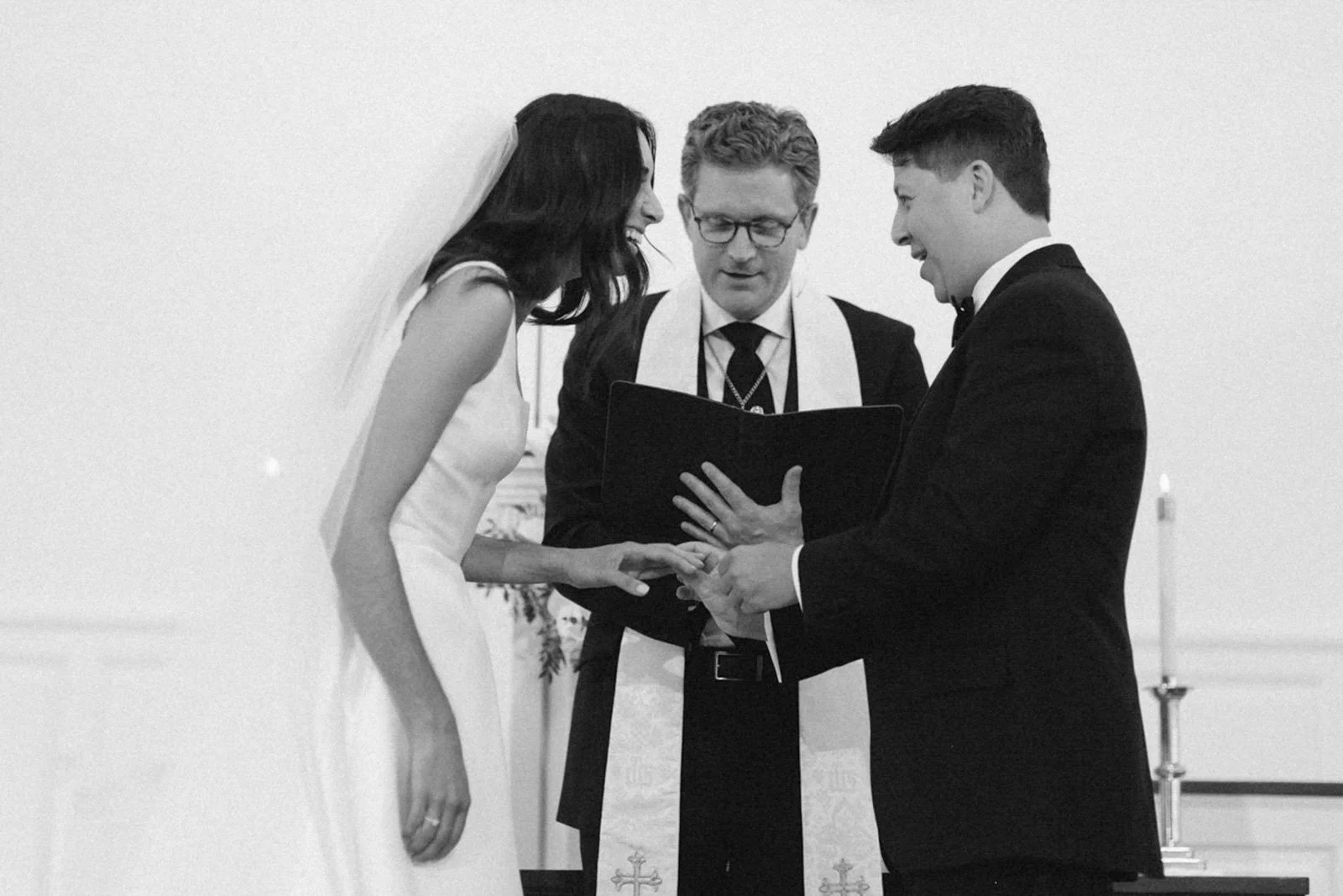 A black and white photo of a wedding ceremony with a bride, groom, and officiant. The bride and groom are holding hands and looking at each other, while the officiant reads from a book.