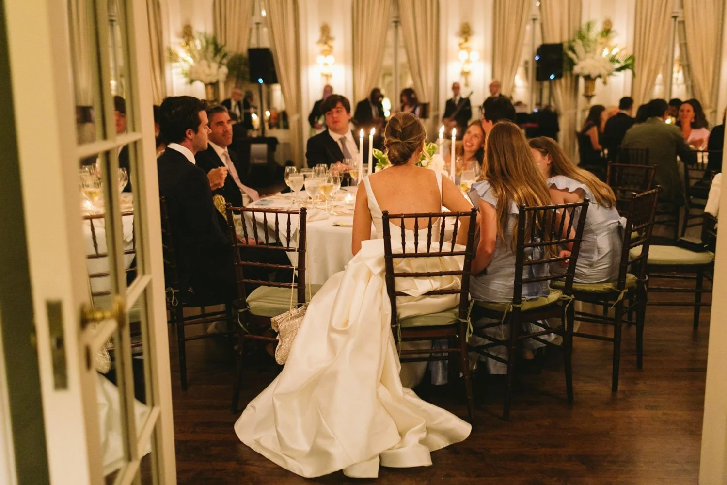 People sitting around a dinner table at a formal event at The Colony Club , likely a wedding reception, in a decorated ballroom with curtains and floral arrangements.