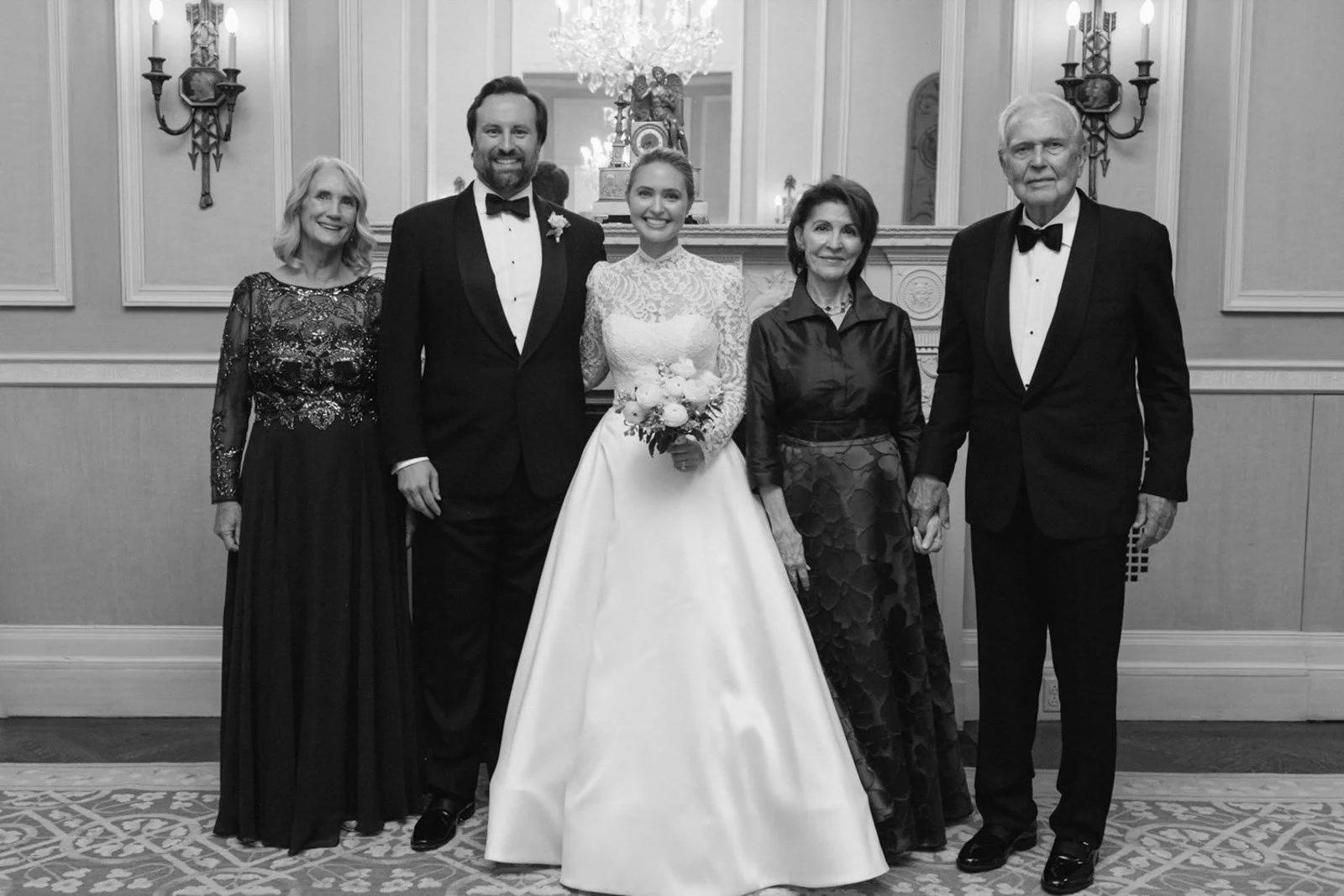 A wedding photo in black and white featuring six people standing together in a formal setting: a bride in a white lace gown holding a bouquet, a groom in a black tuxedo, and four older adults dressed elegantly, possibly family members, in a decorated