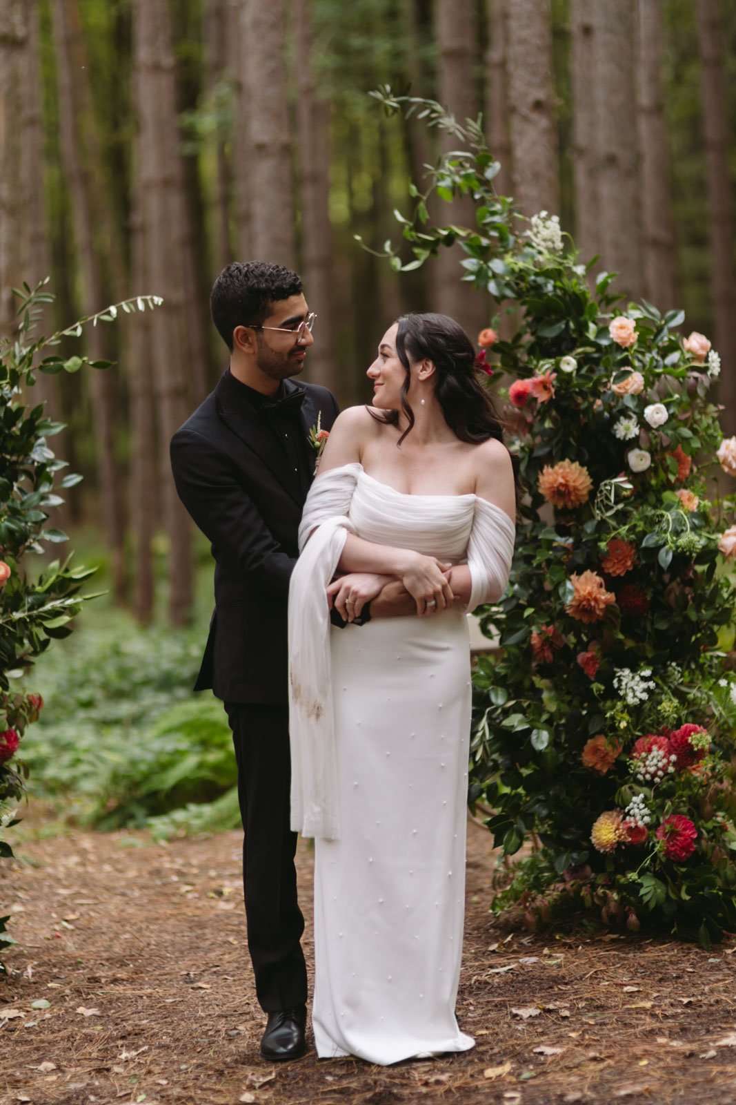 A couple in wedding attire standing in front of a floral archway in a wooded outdoor setting, with the groom looking at the bride and smiling.