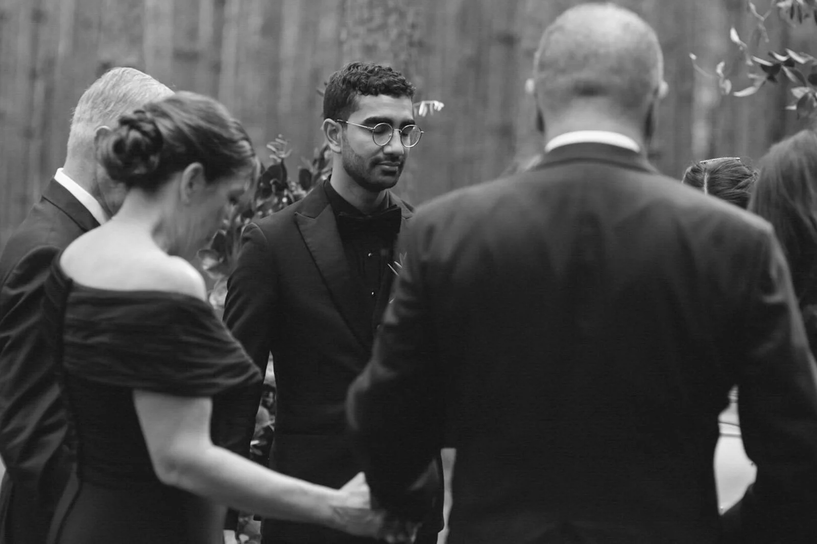 People gathered in a circle, holding hands, at a formal outdoor event. The image is in black and white.