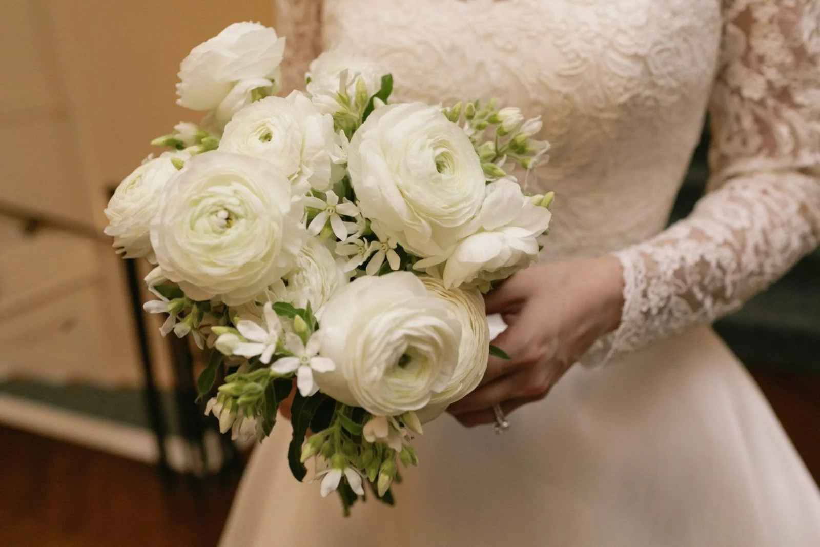 A bride in a lace wedding dress holding a bouquet of white flowers, including ranunculus and small white blooms.