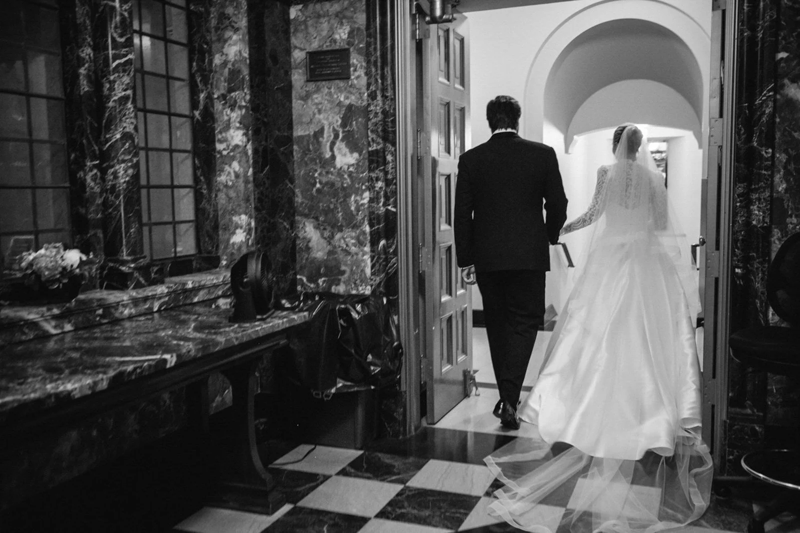 A bride and groom walking hand in hand through a doorway in a church or similar venue, with the bride in a white wedding dress and veil and the groom in a black suit, seen from behind in black and white.