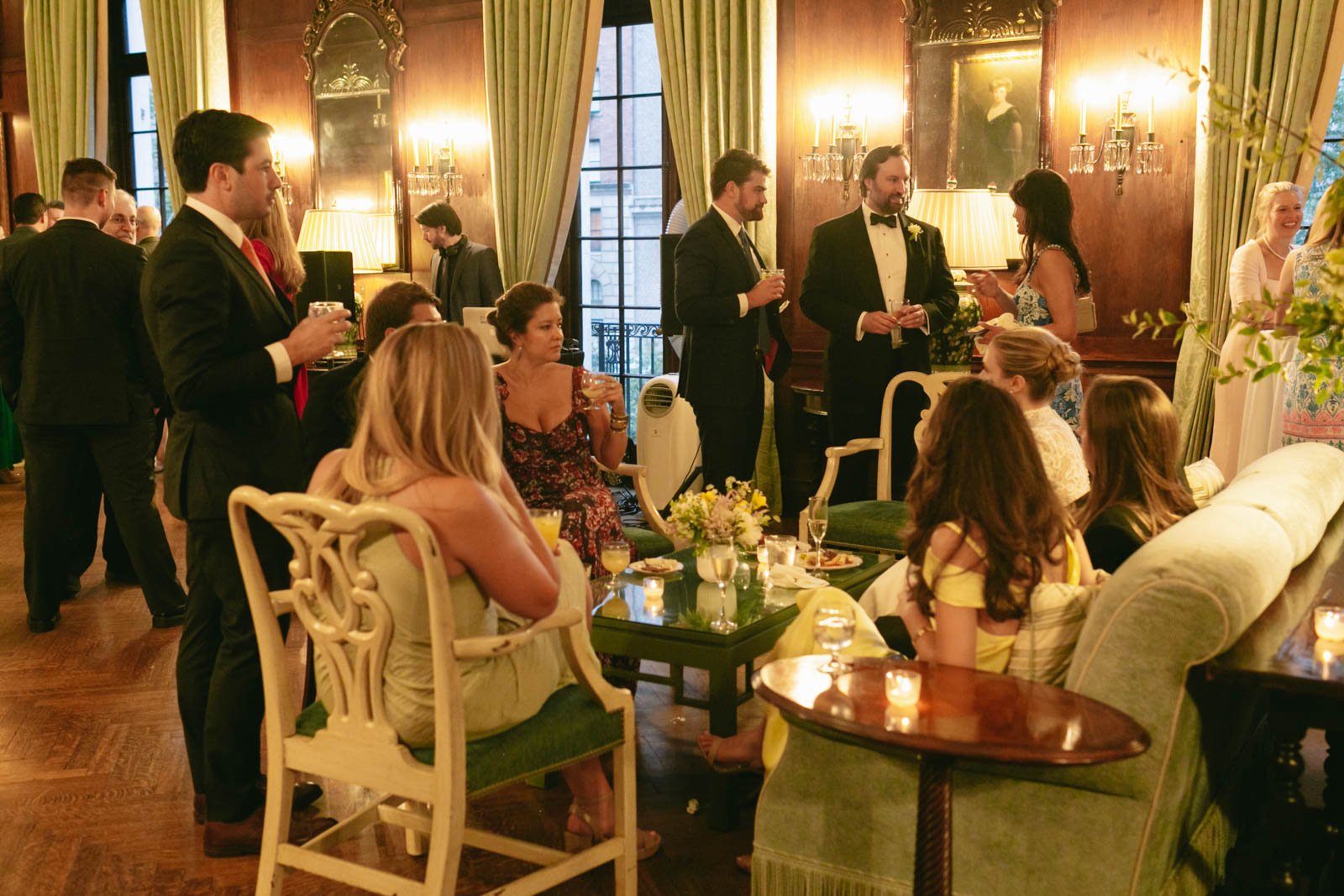 People at a formal gathering in a decorated room with wooden walls, large windows, and curtains, some standing and talking, others sitting with drinks and food, and candles on the tables.