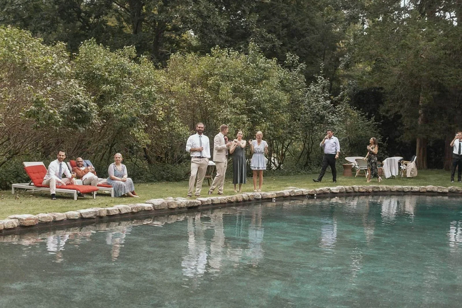 People gathered at a backyard poolside party with trees in the background. Some are sitting on lounge chairs, while others are standing and socializing, holding drinks. Tables and chairs are arranged on the lawn.