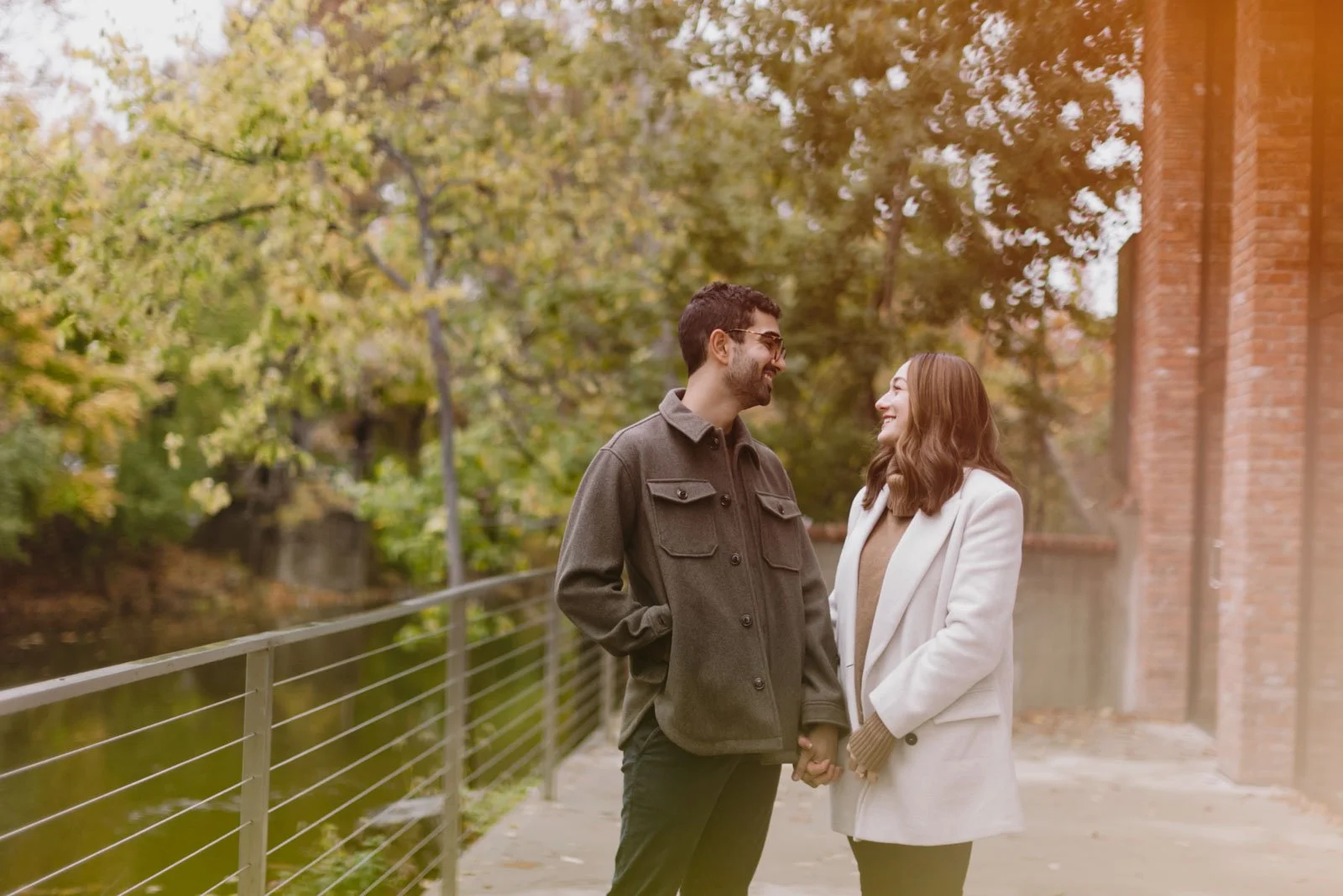 A couple holding hands and smiling at each other outdoors on an autumn day by a river, surrounded by trees with fall foliage.