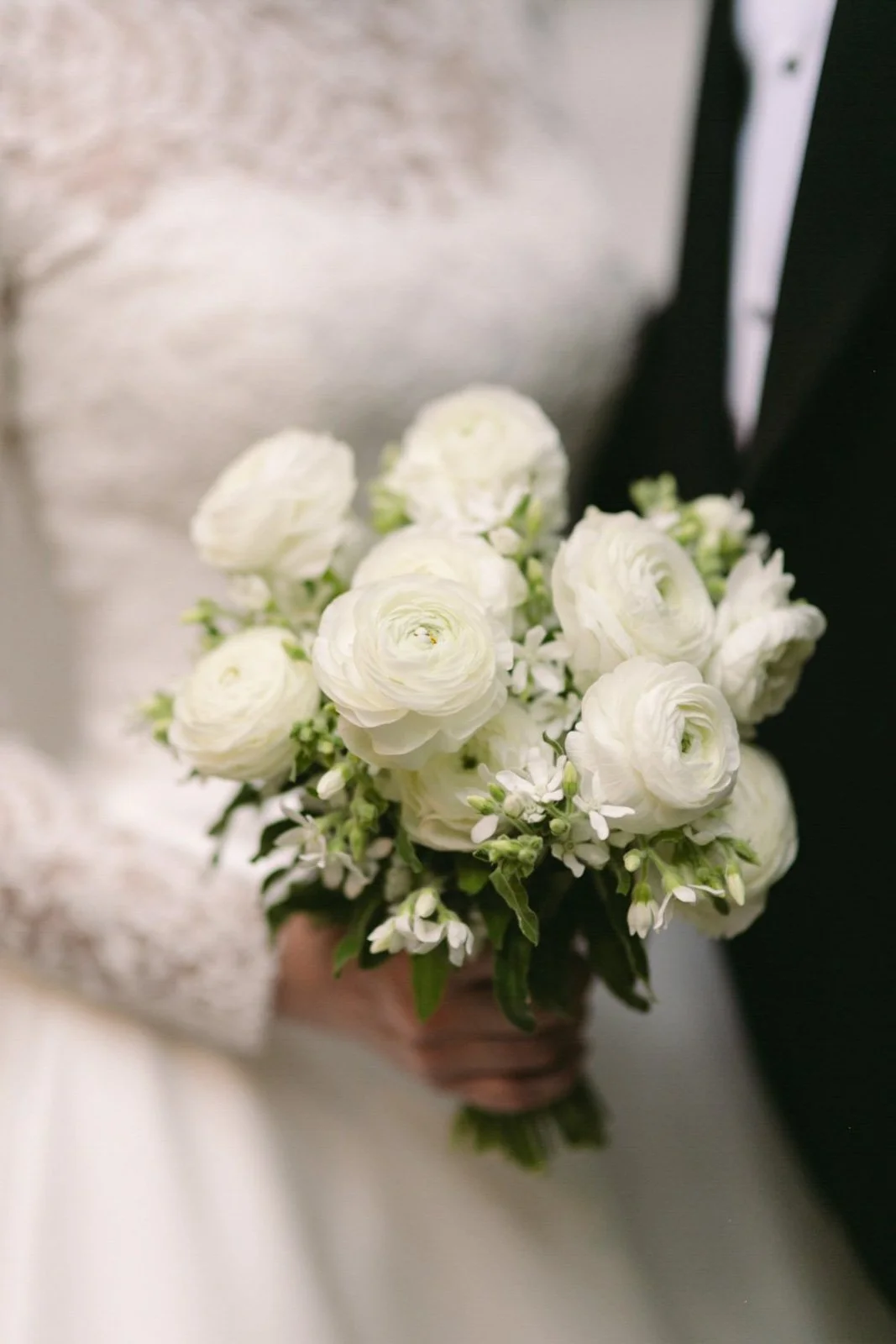 A person holding a bouquet of white flowers, including ranunculus and small accent flowers, in front of a blurred background.