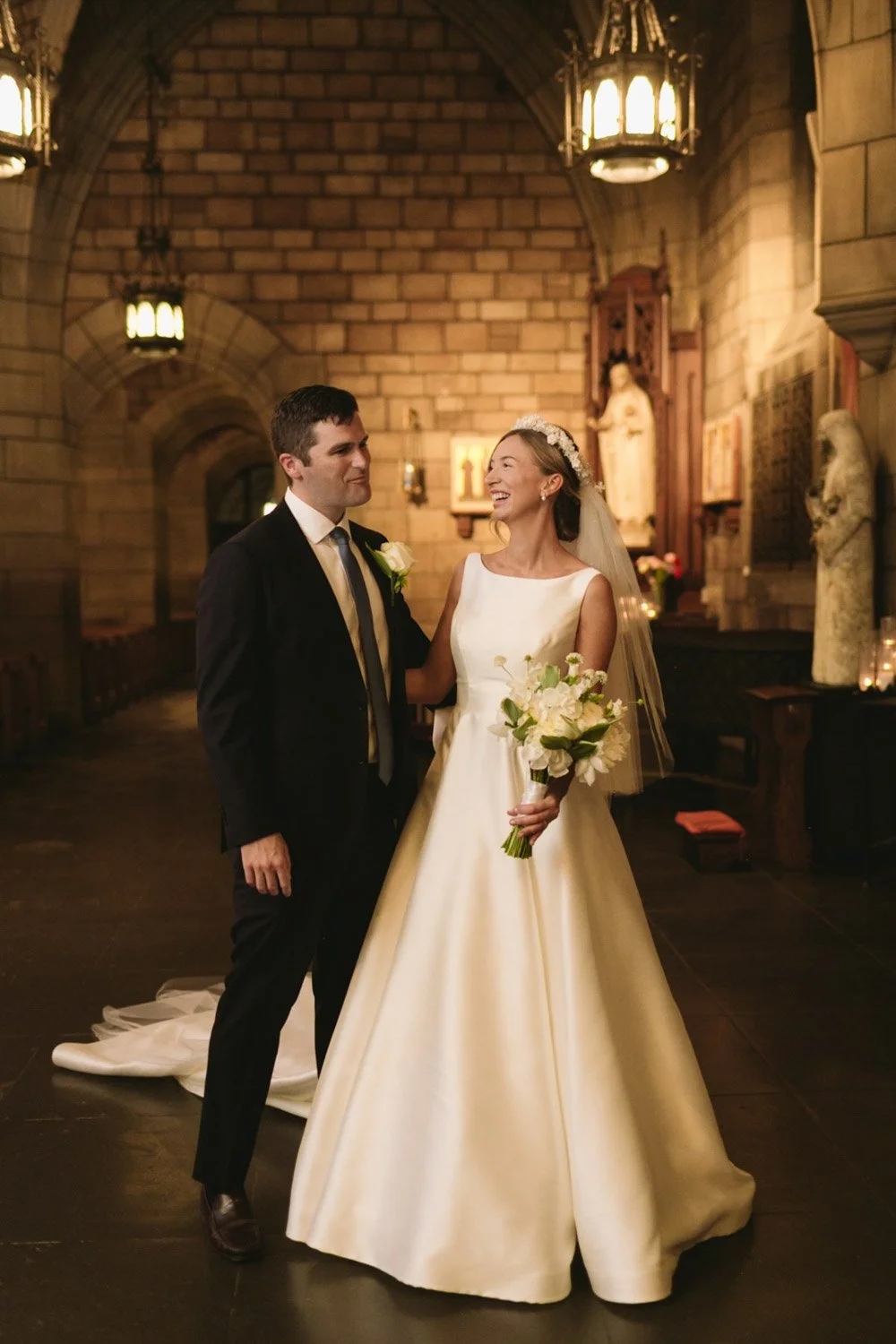 A bride and groom stand inside a church, smiling at each other during their wedding ceremony. The bride wears a white satin gown with a veil and holds a bouquet of white roses. The groom is dressed in a black suit with a white shirt and tie. The chur