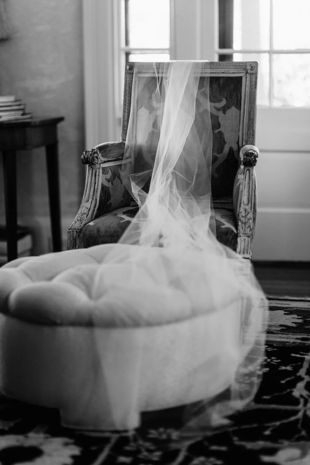 A vintage armchair with a sheer veil draped over the backrest, positioned in a room with a window in the background. There's a tufted ottoman in the foreground and a small table with a stack of books on the side.
