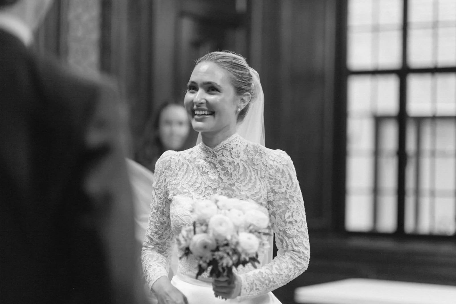A smiling bride in a lace wedding dress holding a bouquet of flowers during a wedding ceremony in a formal setting.
