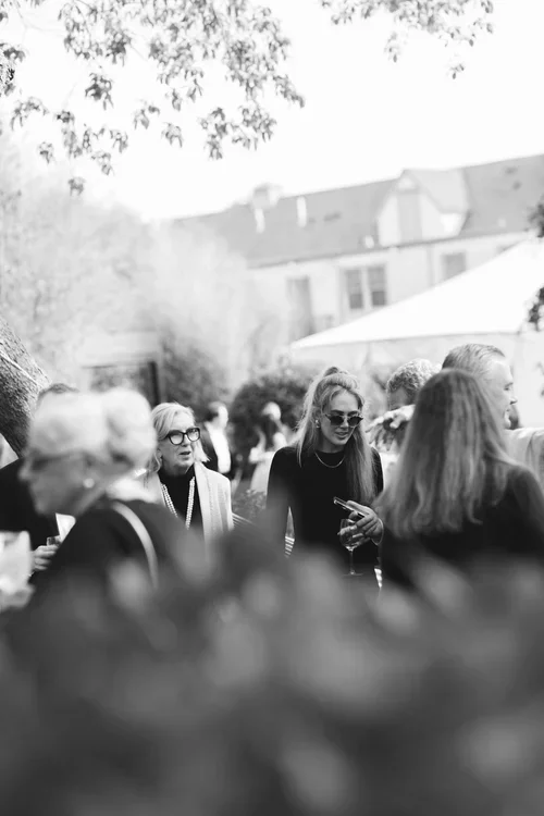 Group of people socializing outdoors at a gathering, with a house and trees in the background.