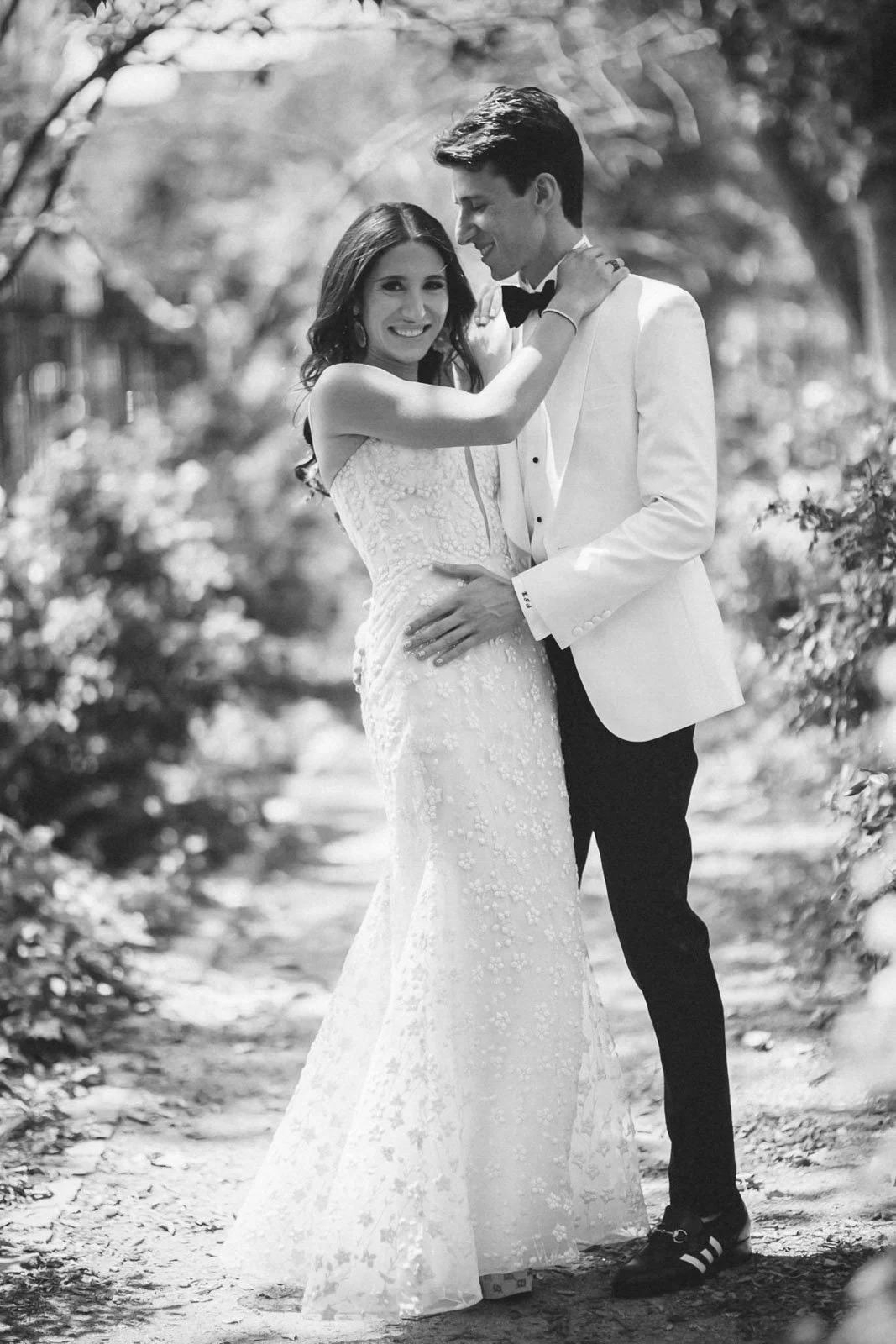 A black and white photo of a smiling bride and groom; the bride in a lace wedding gown, and the groom in a white tuxedo jacket and black bow tie, embracing outdoors.