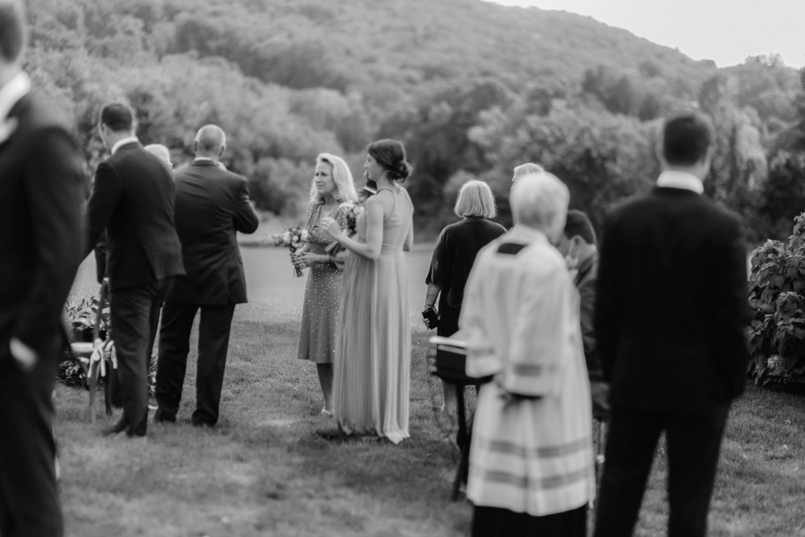 Black and white photo of a group of people outdoors during a wedding or special event, with some standing and talking, a woman in a long dress holding flowers, and a woman with short blonde hair wearing a cardigan and skirt.