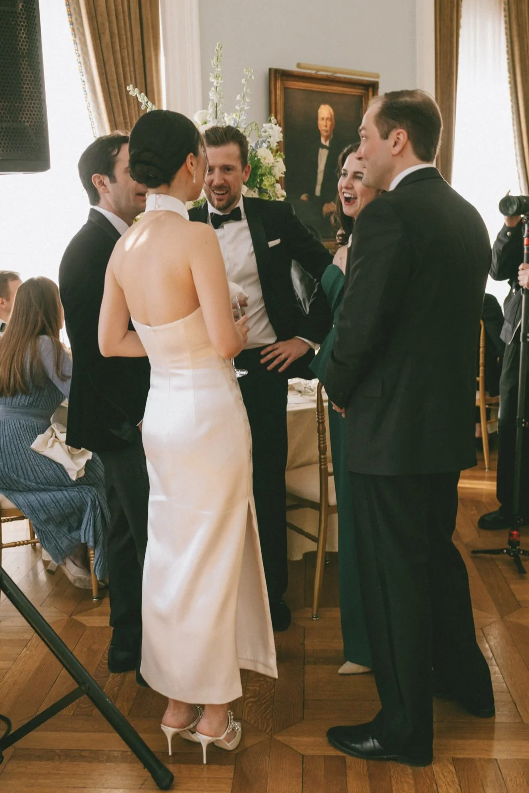 Group of people in formal attire engaged in conversation at a wedding or formal event, with a portrait painting and floral arrangement in the background.