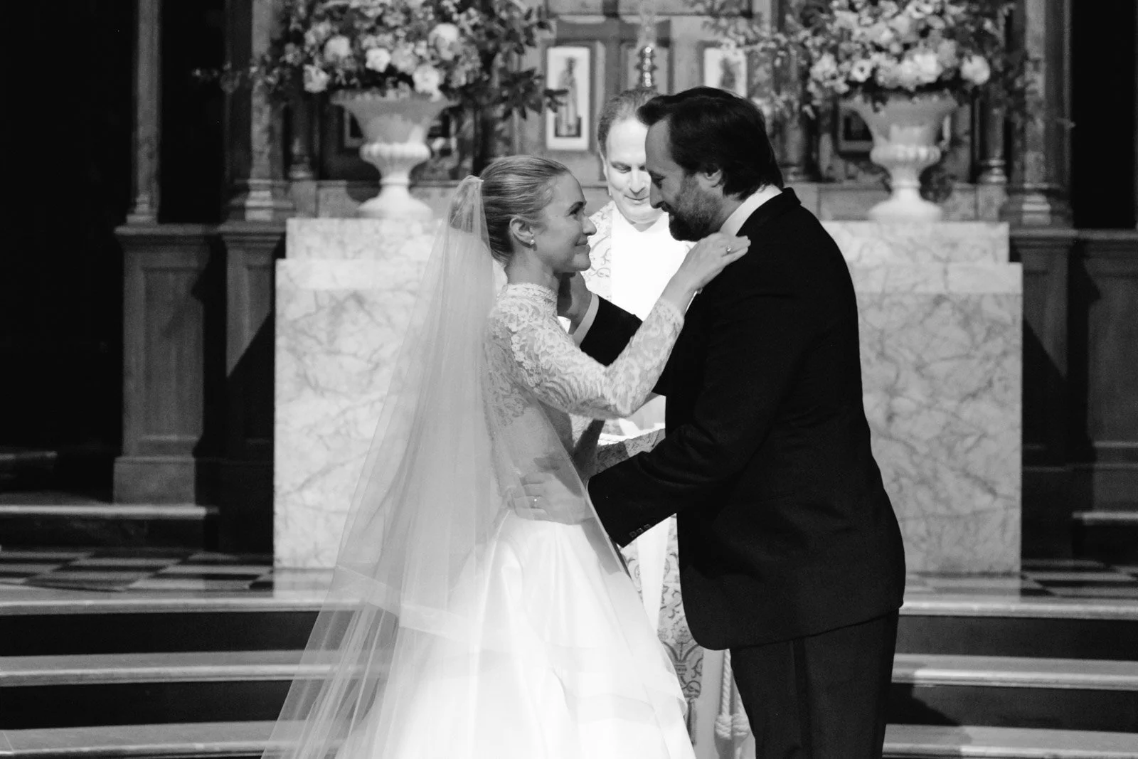 Black and white photo of a bride and groom during a wedding ceremony, standing close and gazing at each other, with an officiant in the background.