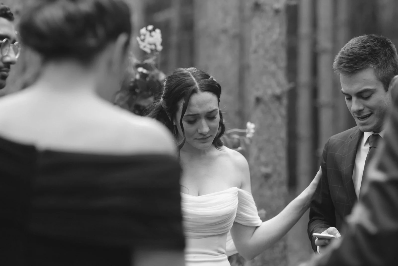 Black-and-white photo of a wedding ceremony with a woman in an off-shoulder dress and a man in a suit, surrounded by onlookers, outdoors near trees.