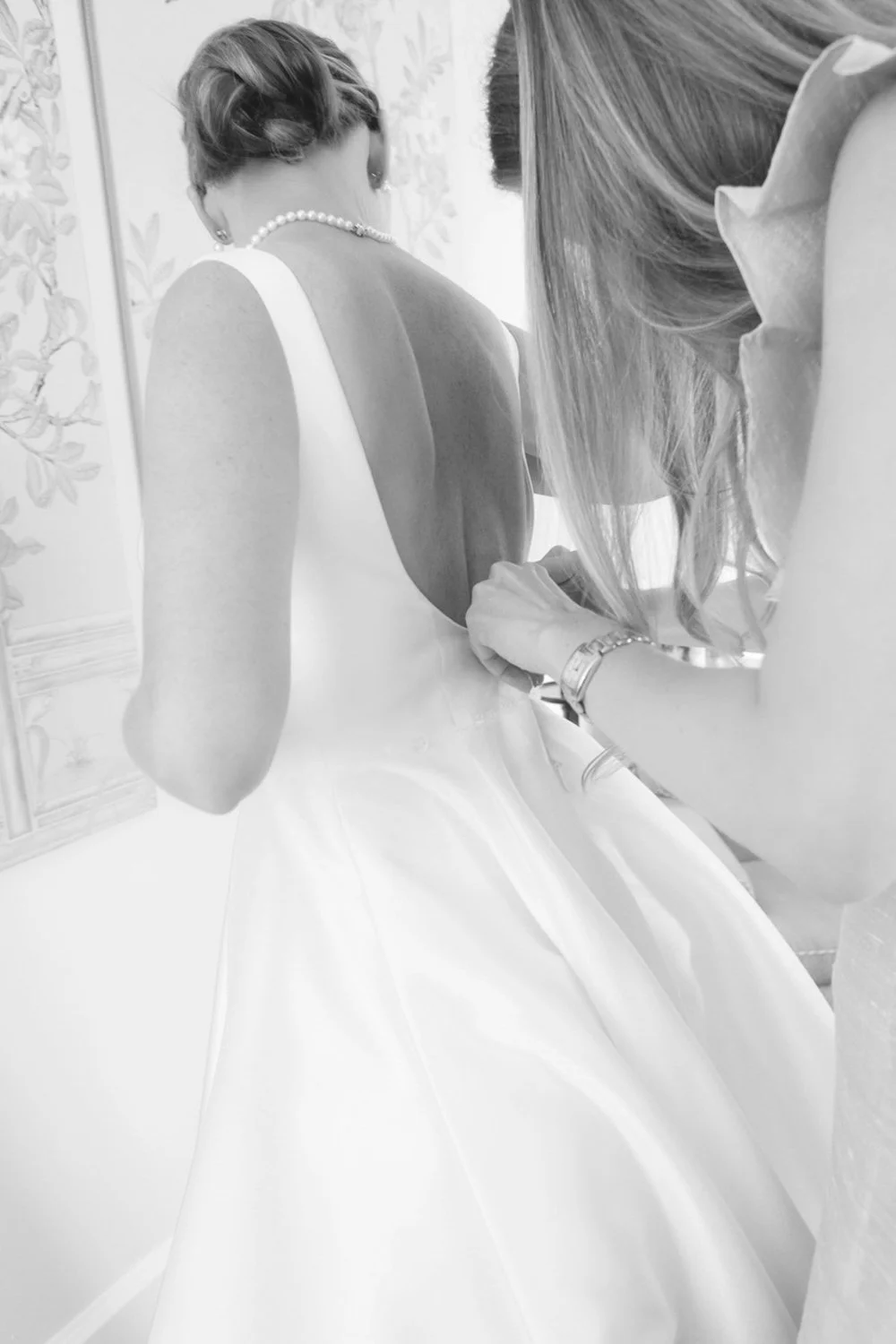 A woman in a white wedding dress getting dressed, with assistance from another woman, in a softly lit room.